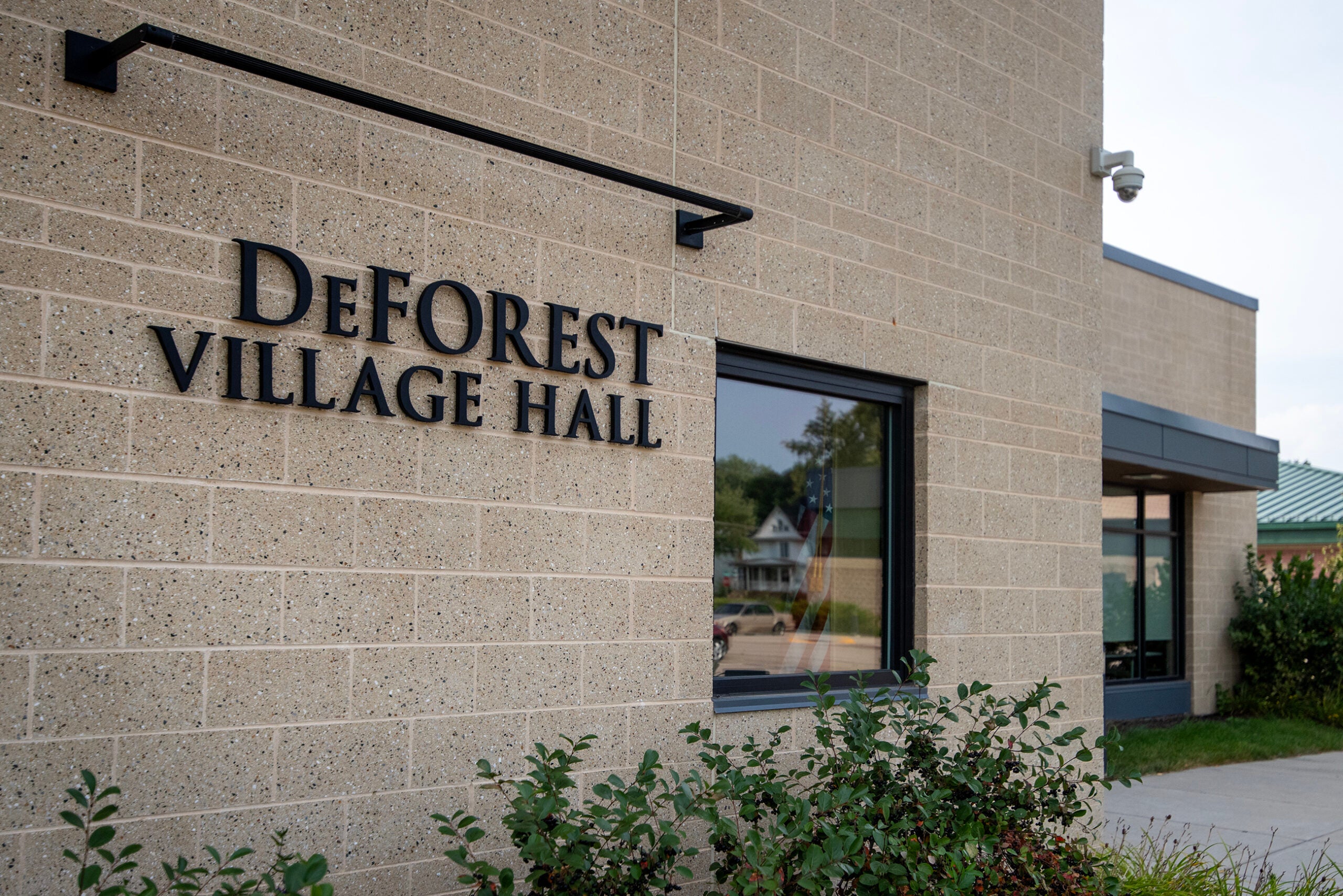 Exterior of DeForest Village Hall building with tan brick walls, large windows, and a sign displaying the buildings name. Shrubs are visible in the foreground.