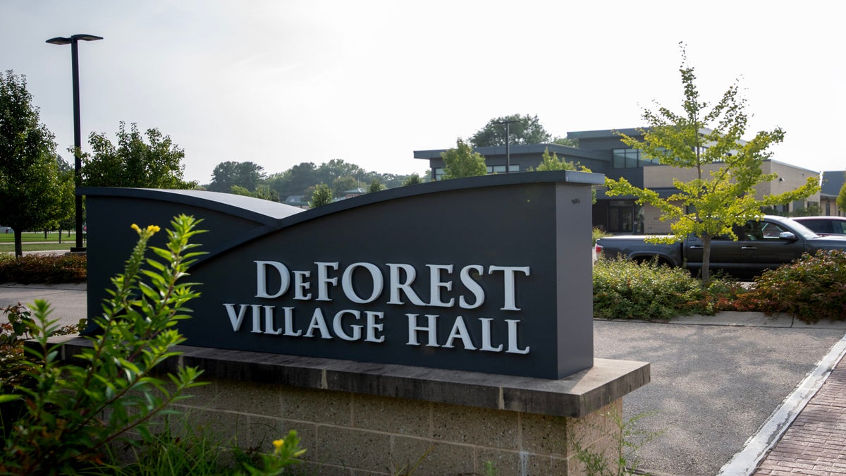 A sign reading DeForest Village Hall stands in front of a modern building, with trees, parked cars, and a sidewalk visible in the background.