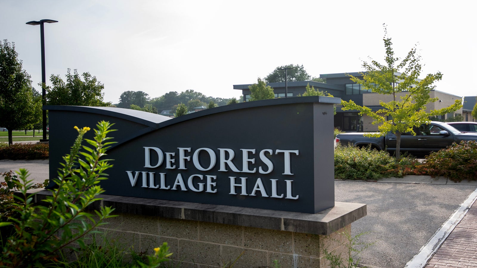 A sign reading DeForest Village Hall stands in front of a modern building, with trees, parked cars, and a sidewalk visible in the background.
