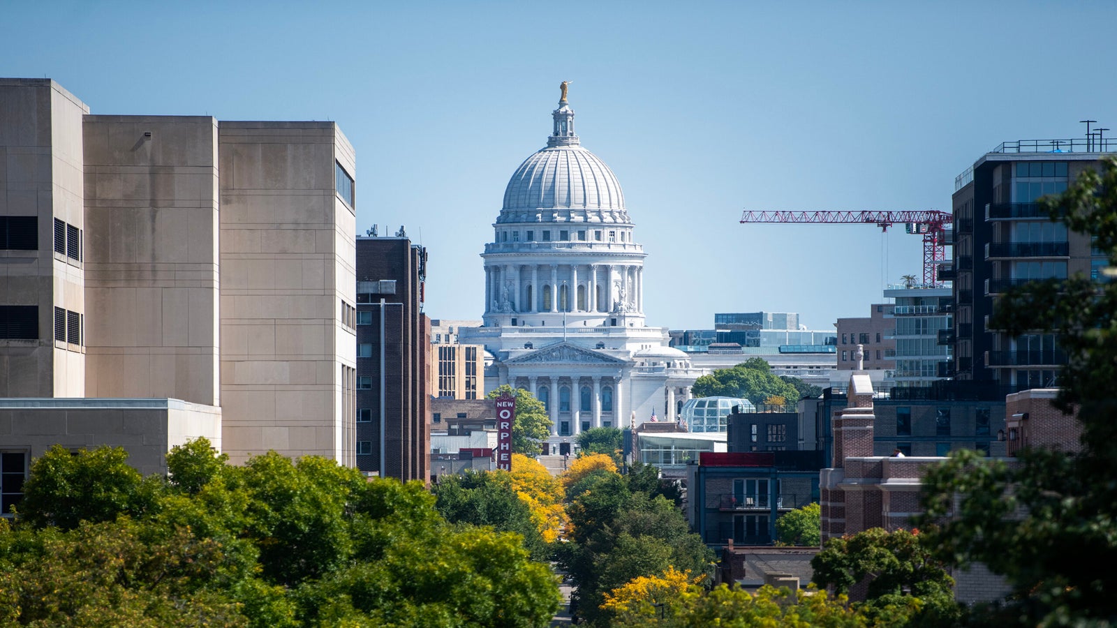A view of the Wisconsin State Capitol building centered between city buildings, with trees and a construction crane in the foreground.