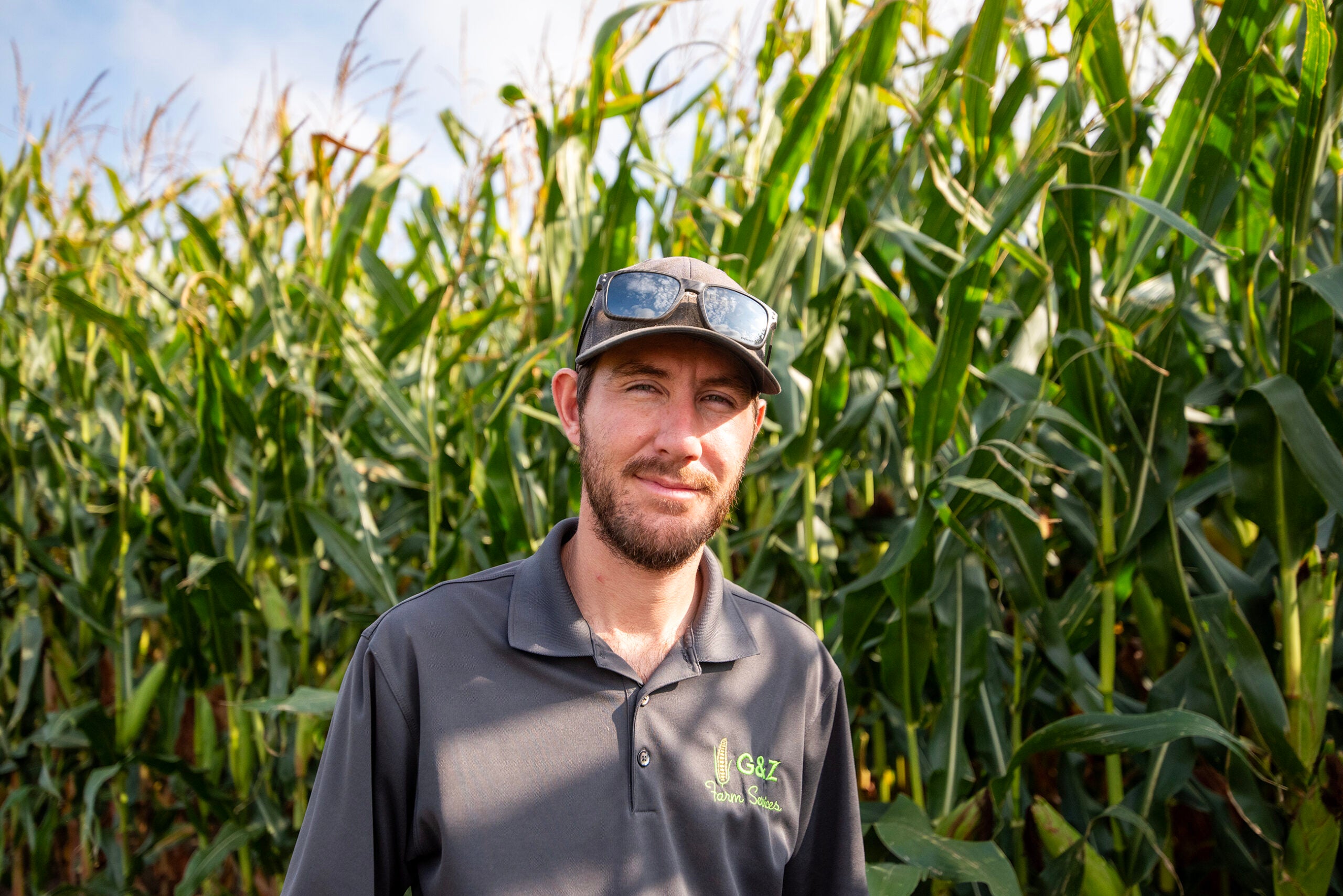 A man wearing a dark polo shirt and cap stands in front of tall corn plants under a blue sky. Sunglasses rest on his hat.