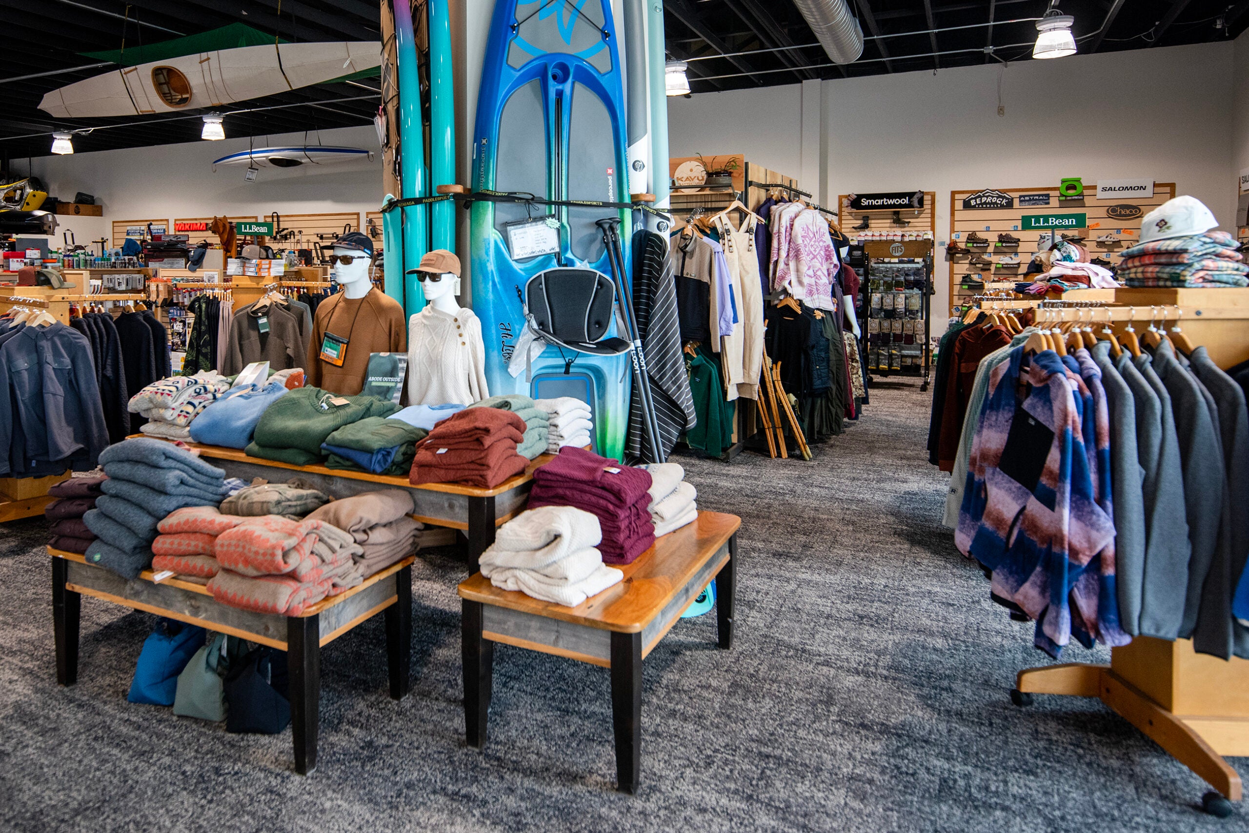 Clothing store interior with folded sweaters on tables, mannequins dressed in casual wear, and various apparel hanging on racks. Surfboards and paddles are displayed in the background.