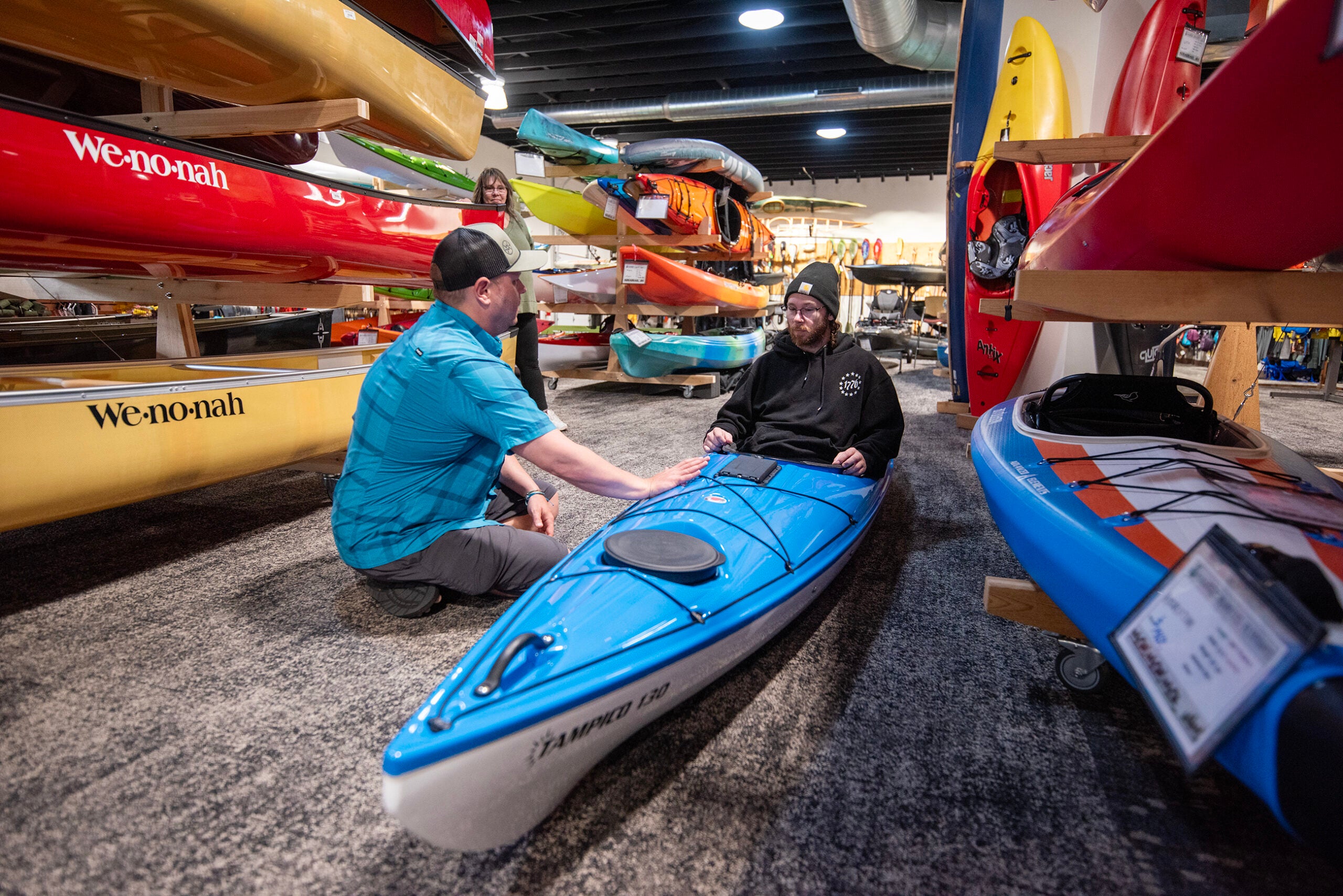 Two men are inside a kayak shop; one sits in a blue kayak on the floor while the other kneels beside him, surrounded by various colorful kayaks on display.