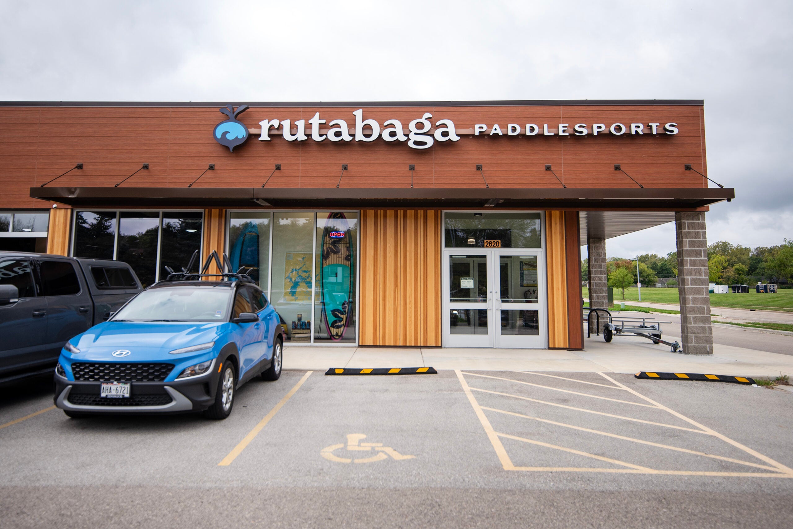 A blue car is parked outside Rutabaga Paddlesports, a store with a wooden facade and glass entrance. A handicapped parking space is in the foreground.