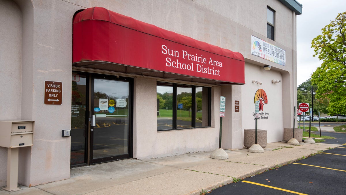 Entrance to the Sun Prairie Area School District building with a red awning, glass doors, and a visitor parking sign visible.