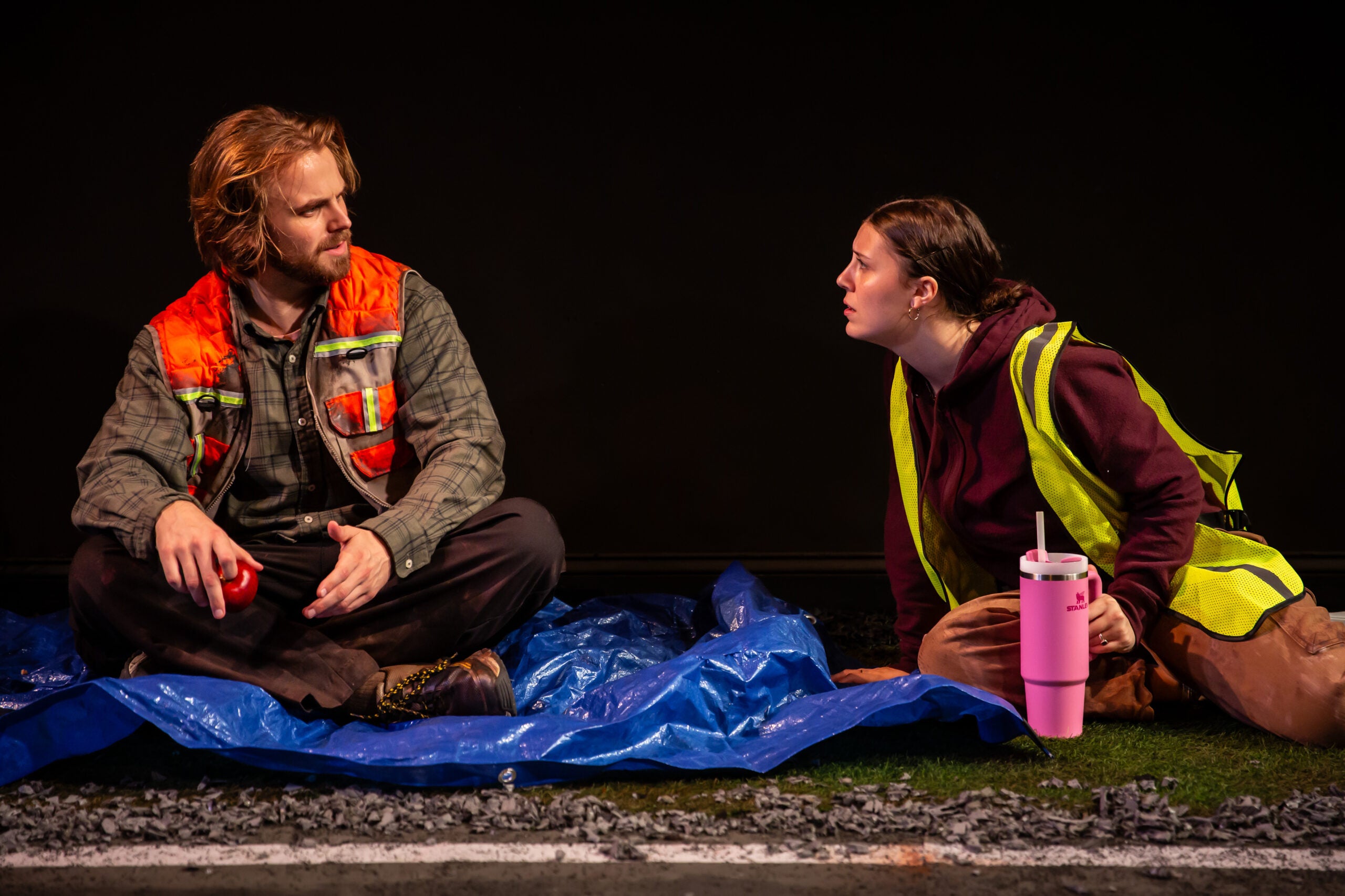 Two people in work vests sit on a blue tarp, talking; one holds a cup, the other has a large pink tumbler, with gravel and grass visible at their feet.