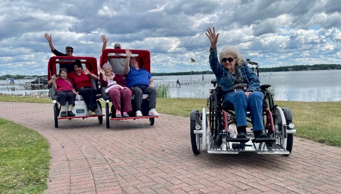 Four people sit in two rickshaw-style bikes, and one woman rides a modified wheelchair bike on a lakeside path; all are waving and smiling under a cloudy sky.
