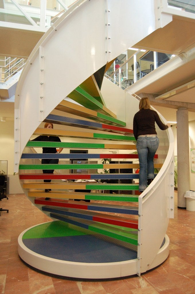 A person ascends a modern spiral staircase with colorful steps inside a bright, open building. Another person is visible on the lower floor.