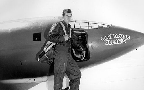 A man in a flight suit stands beside the open cockpit of an aircraft labeled Glamorous Glennis.