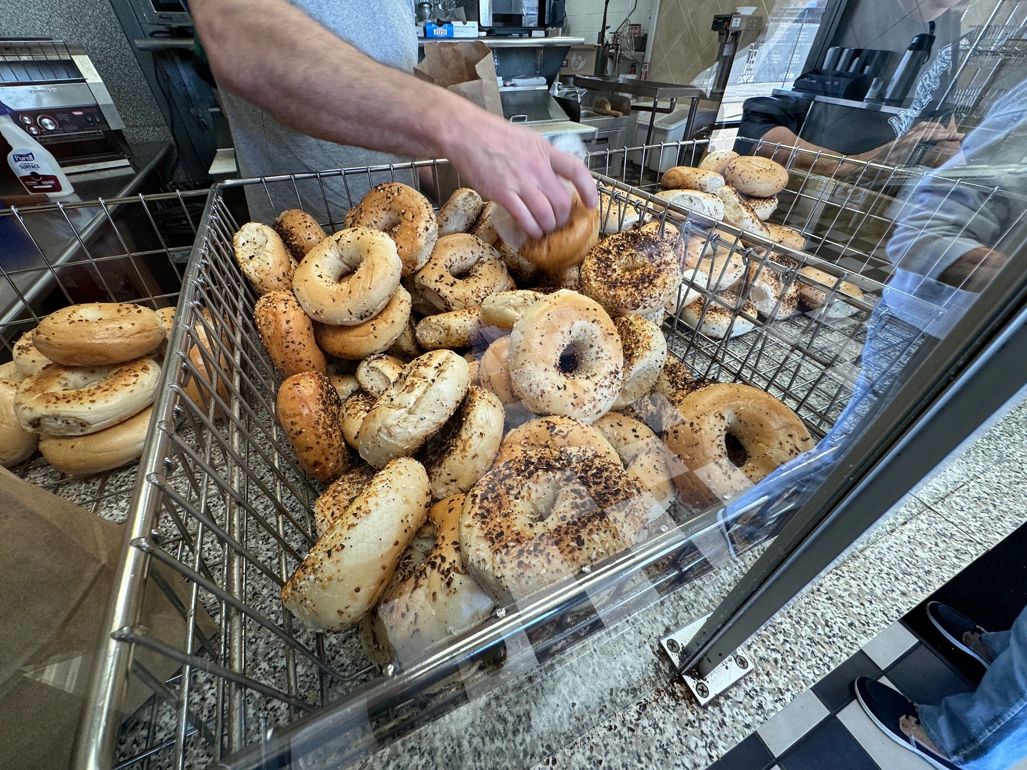 A person selects a bagel from a large metal bin filled with assorted bagels at a bakery or bagel shop.