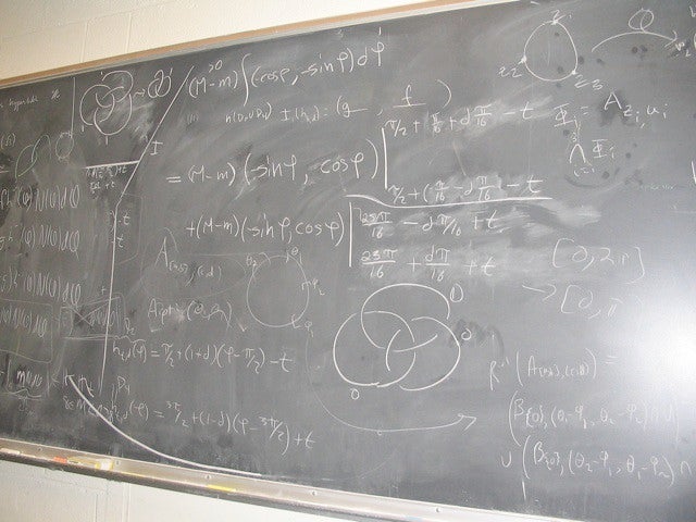 A chalkboard filled with mathematical equations, formulas, and diagrams, including circles, graphs, and various algebraic expressions written in white chalk.