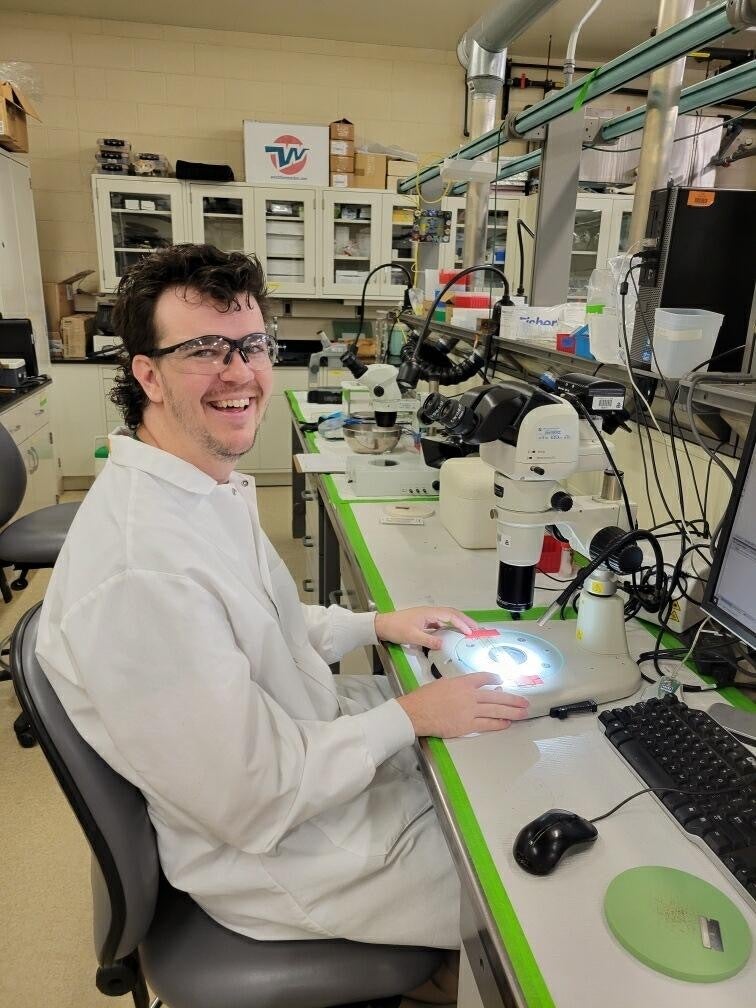 A person wearing a lab coat and safety glasses sits at a laboratory workstation, smiling at the camera, with microscopes and lab equipment on the desk.