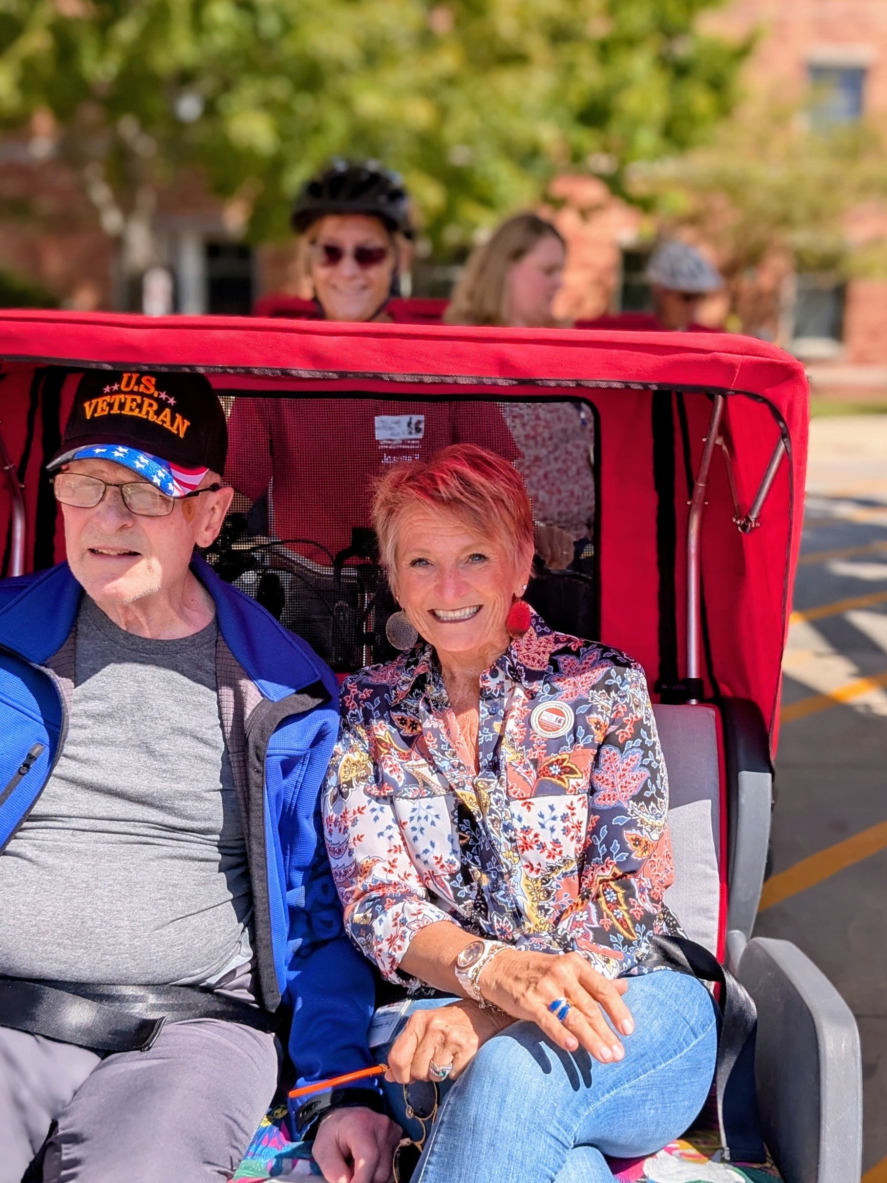 An older man wearing a U.S. Veteran cap sits next to a smiling woman in a floral shirt on a red rickshaw, with two people in the background.