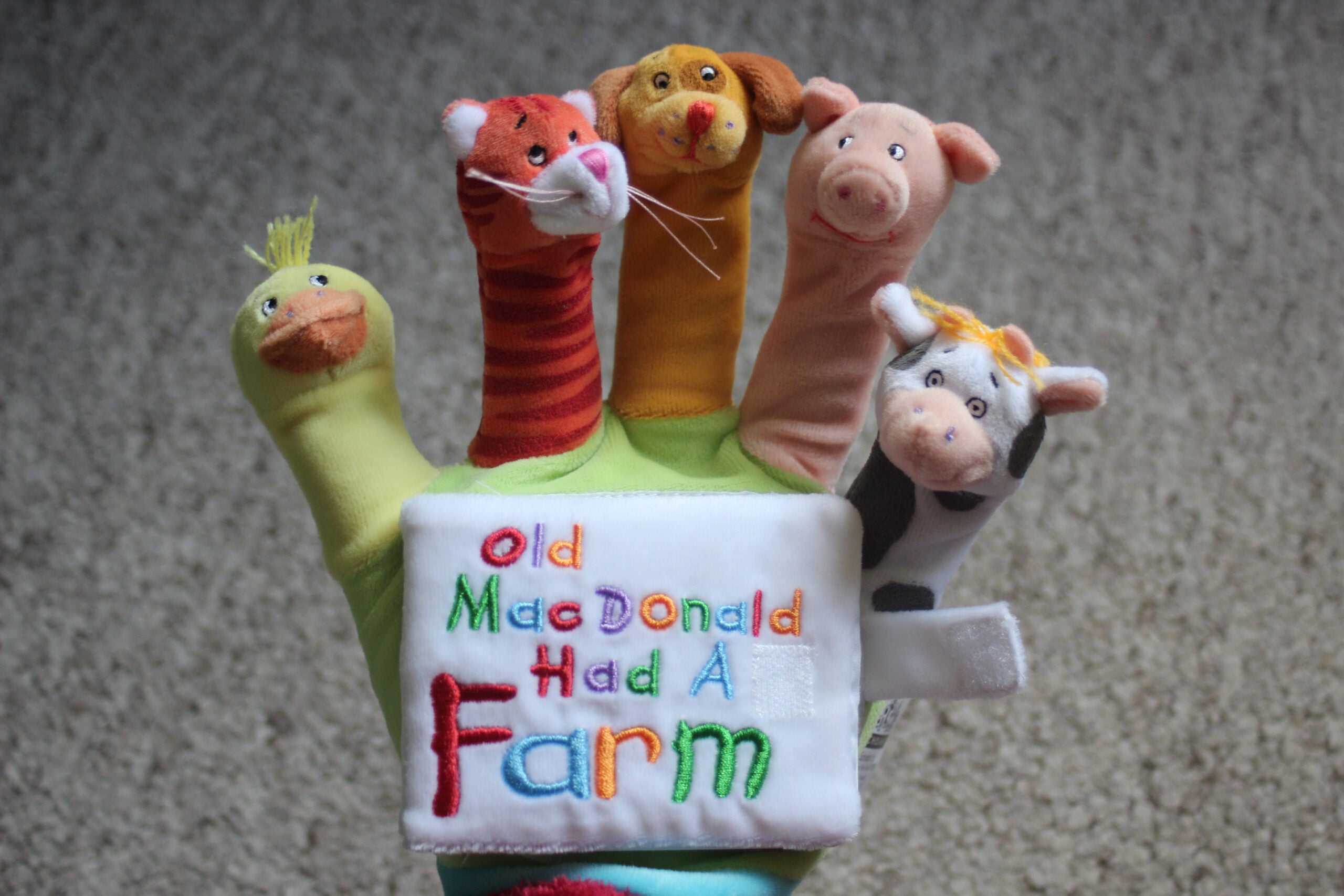 A plush glove puppet with animal heads on each finger and the words Old MacDonald Had A Farm embroidered on the palm, lying on a carpeted surface.