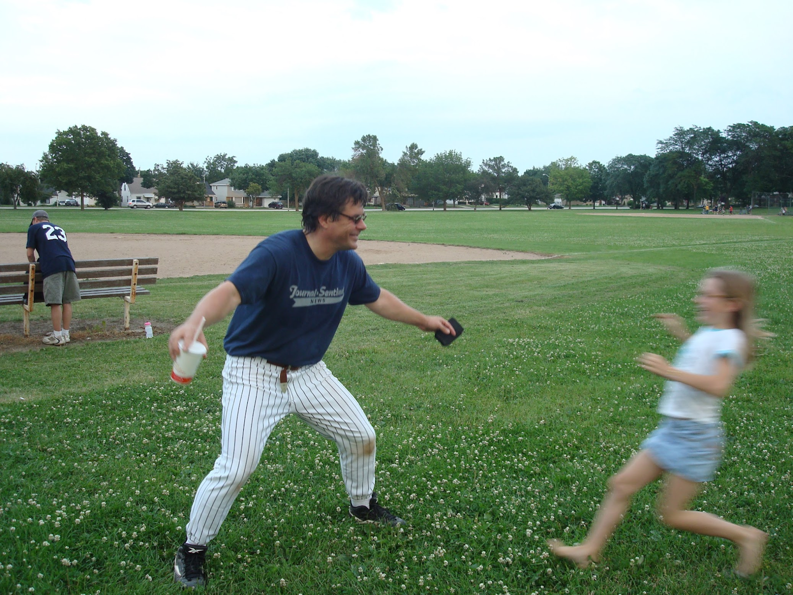 An adult in striped pants and a blue shirt stands on grass with arms outstretched toward a running child; a baseball field and another person are in the background.