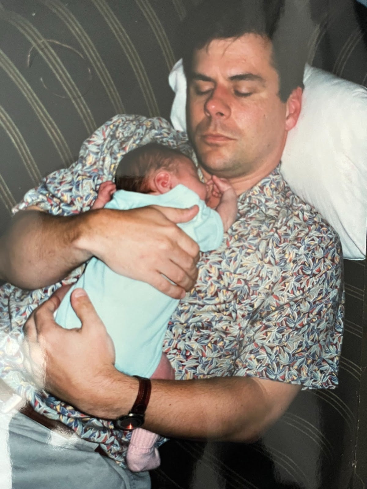 An adult man lying on a couch with his eyes closed, holding a sleeping baby dressed in light blue clothing on his chest.