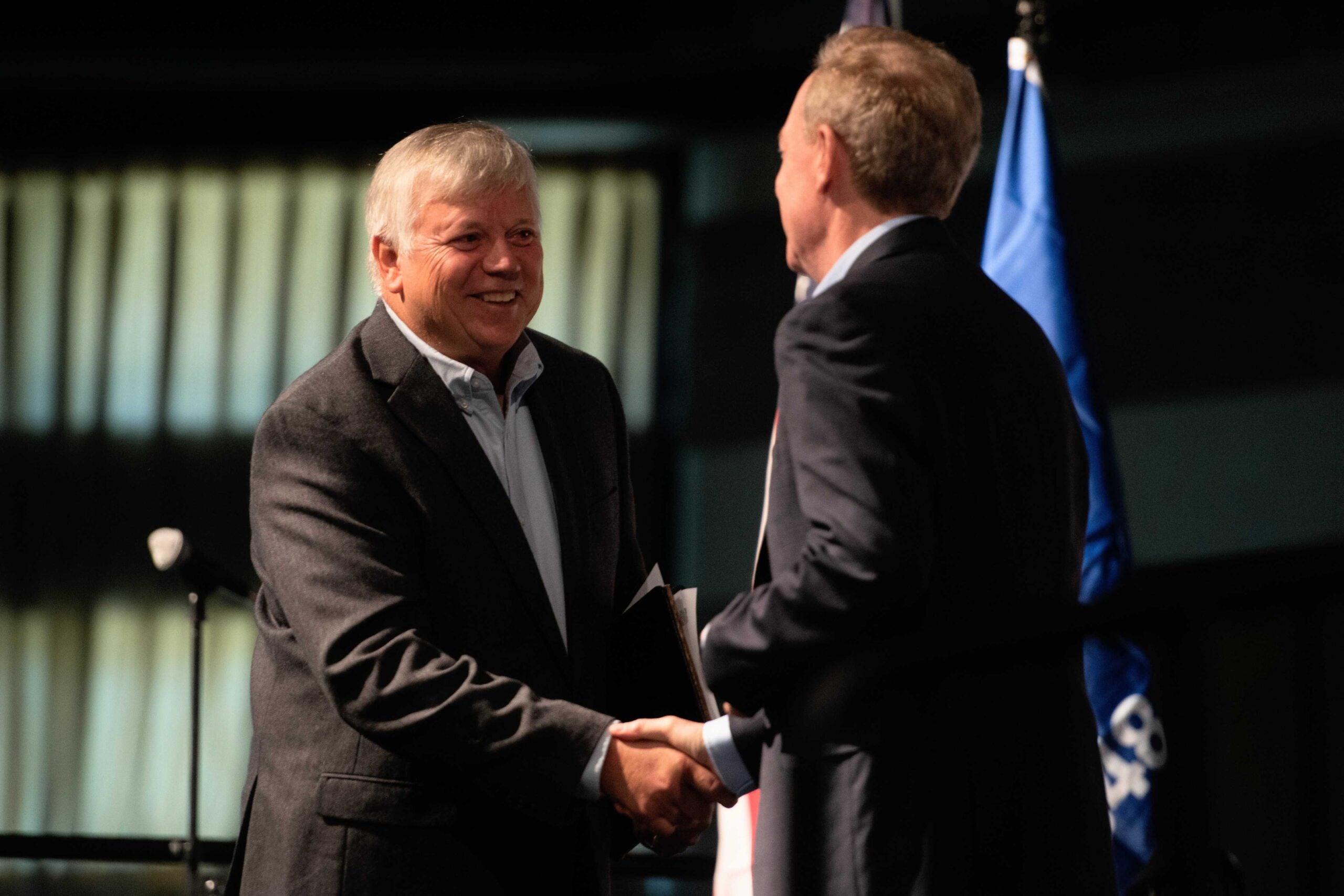 Two men in suits shake hands onstage in front of flags and curtains, one holding a folder and smiling at the other.