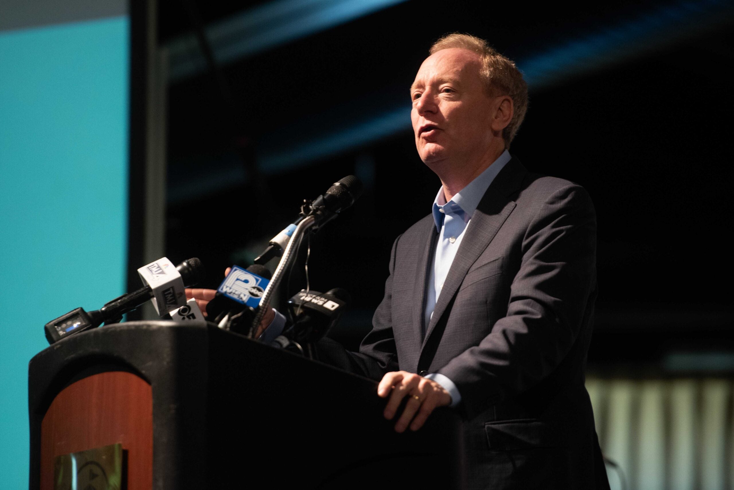 A man in a suit speaks at a podium with several microphones during an indoor event.