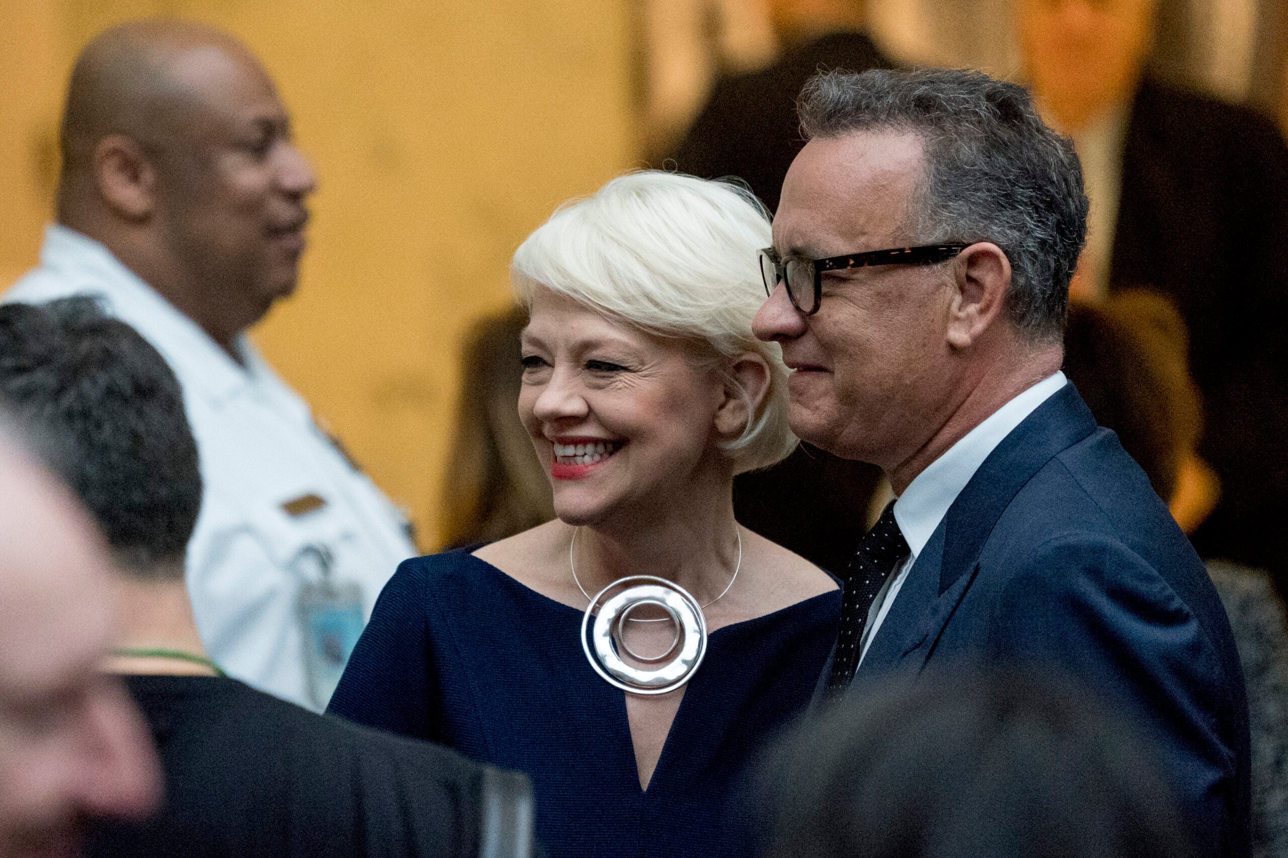 A woman with short blonde hair and a man in a suit and glasses stand together at a formal event, smiling, with other people visible in the background.