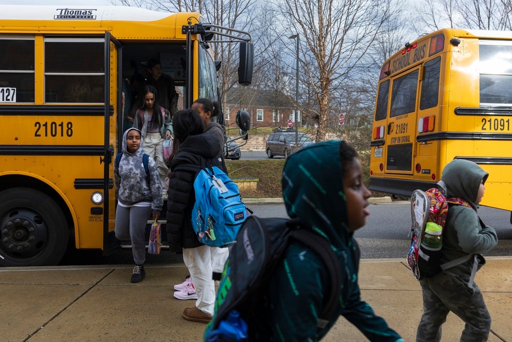 Children exit a yellow school bus while others walk on the sidewalk outside, with another school bus parked nearby on a cloudy day.