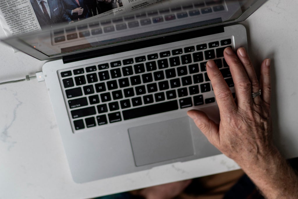 A hand with a ring rests on the keyboard of a laptop, seen from above. Part of a newspaper is visible above the keyboard.