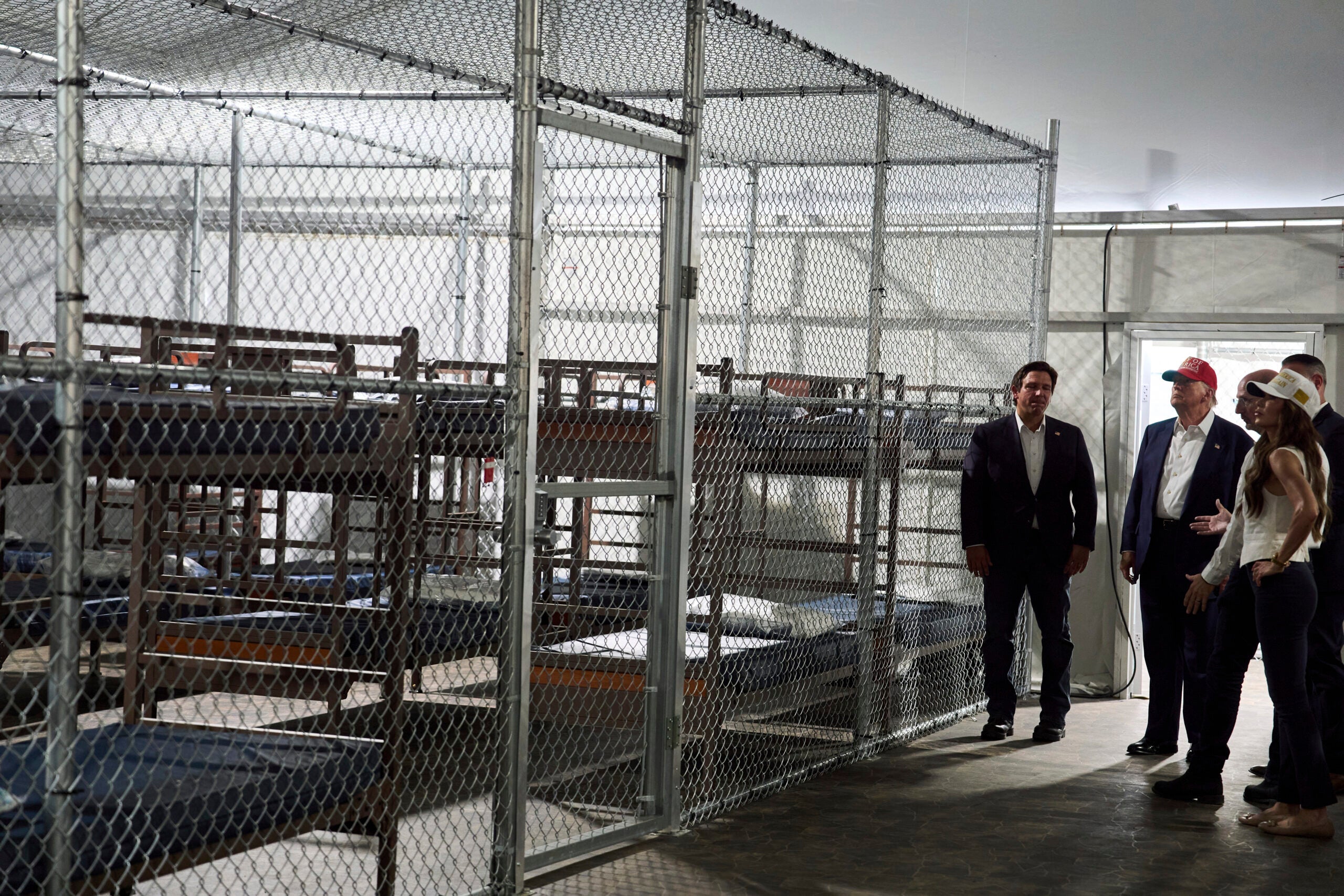 Several people stand near a fenced-off area containing rows of metal bunk beds inside a large, industrial-style building.