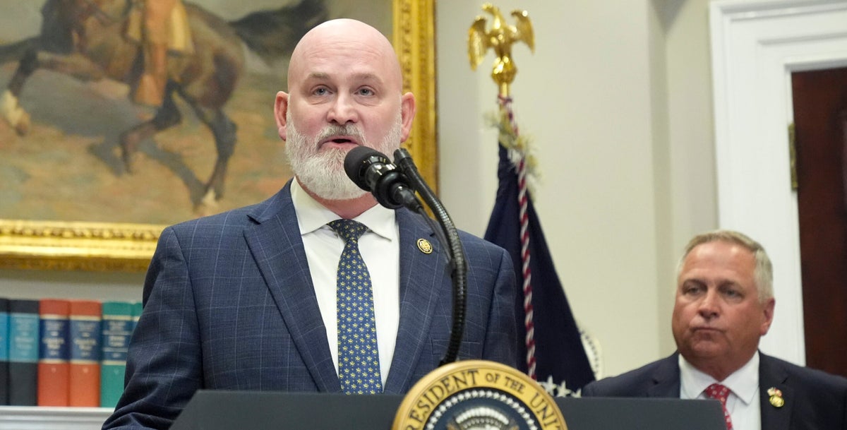 A man in a suit speaks at a podium with the presidential seal, while another man stands behind him in a room with a painting, books, and a U.S. flag.
