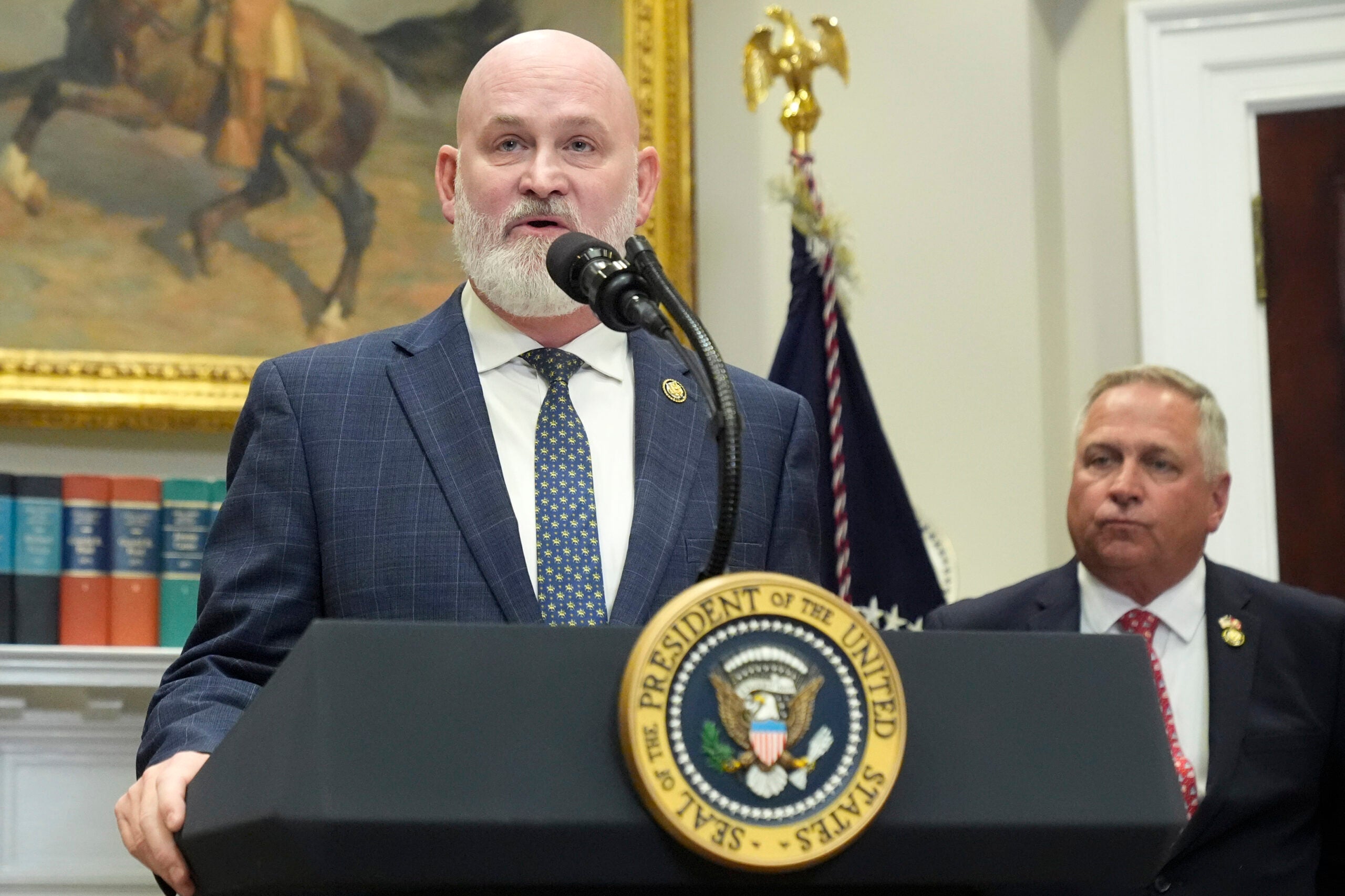 A man in a suit speaks at a podium with the presidential seal, while another man stands behind him in a room with a painting, books, and a U.S. flag.