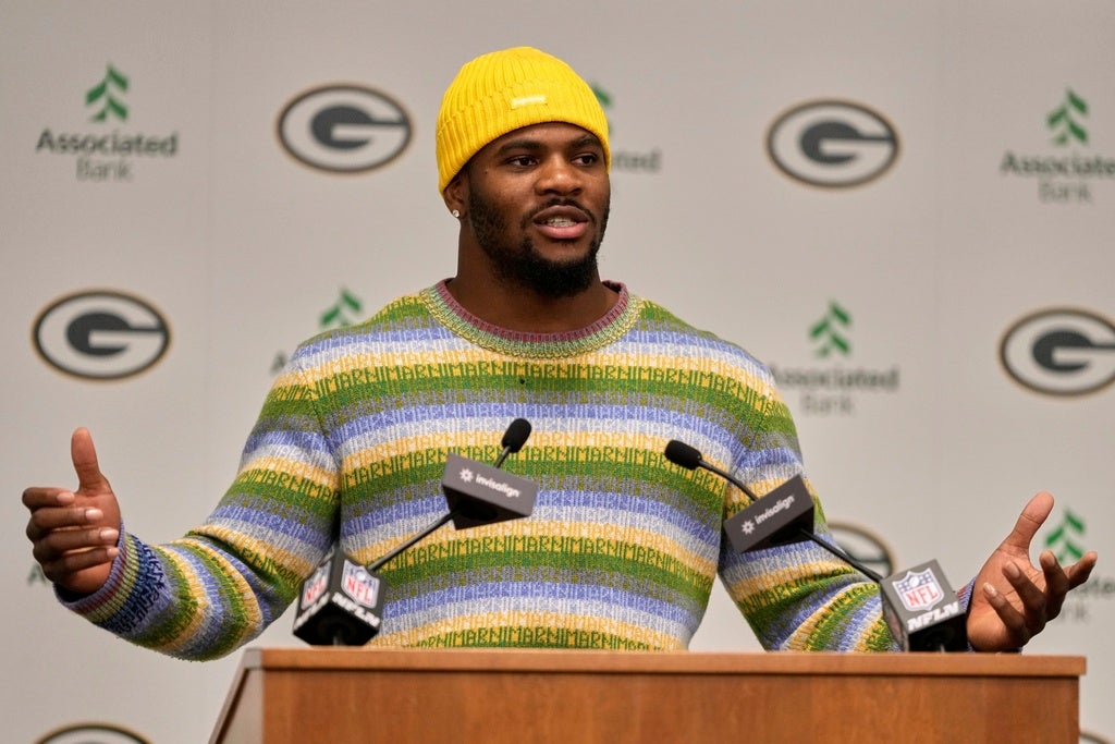 A man in a yellow beanie and striped sweater speaks at an NFL press conference podium with Green Bay Packers and Associated Bank logos in the background.