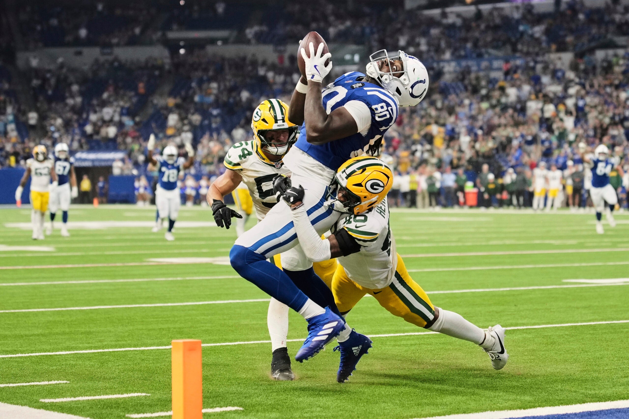 A football player in a blue uniform catches a pass in the end zone while being tackled by two defenders in yellow and green uniforms during a game.