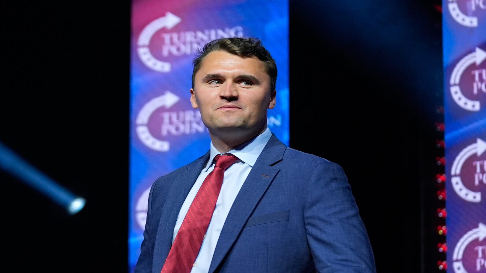 A man in a blue suit and red tie stands on stage in front of Turning Point USA banners, with stage lights in the background.