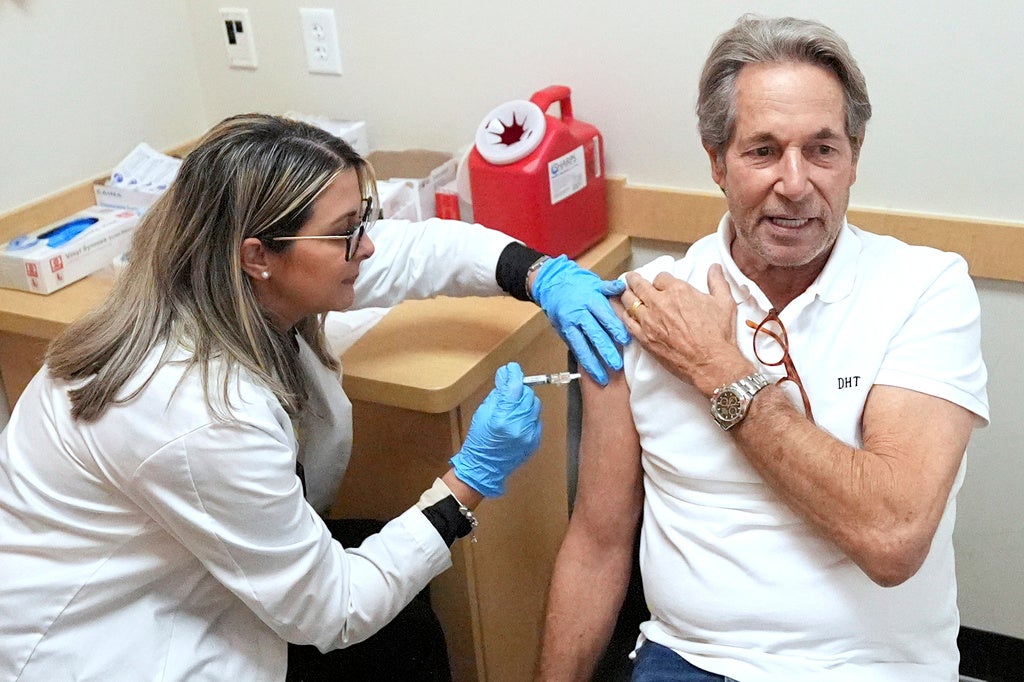 A healthcare worker wearing gloves administers a vaccine injection into the upper arm of an older man seated in a clinic room.