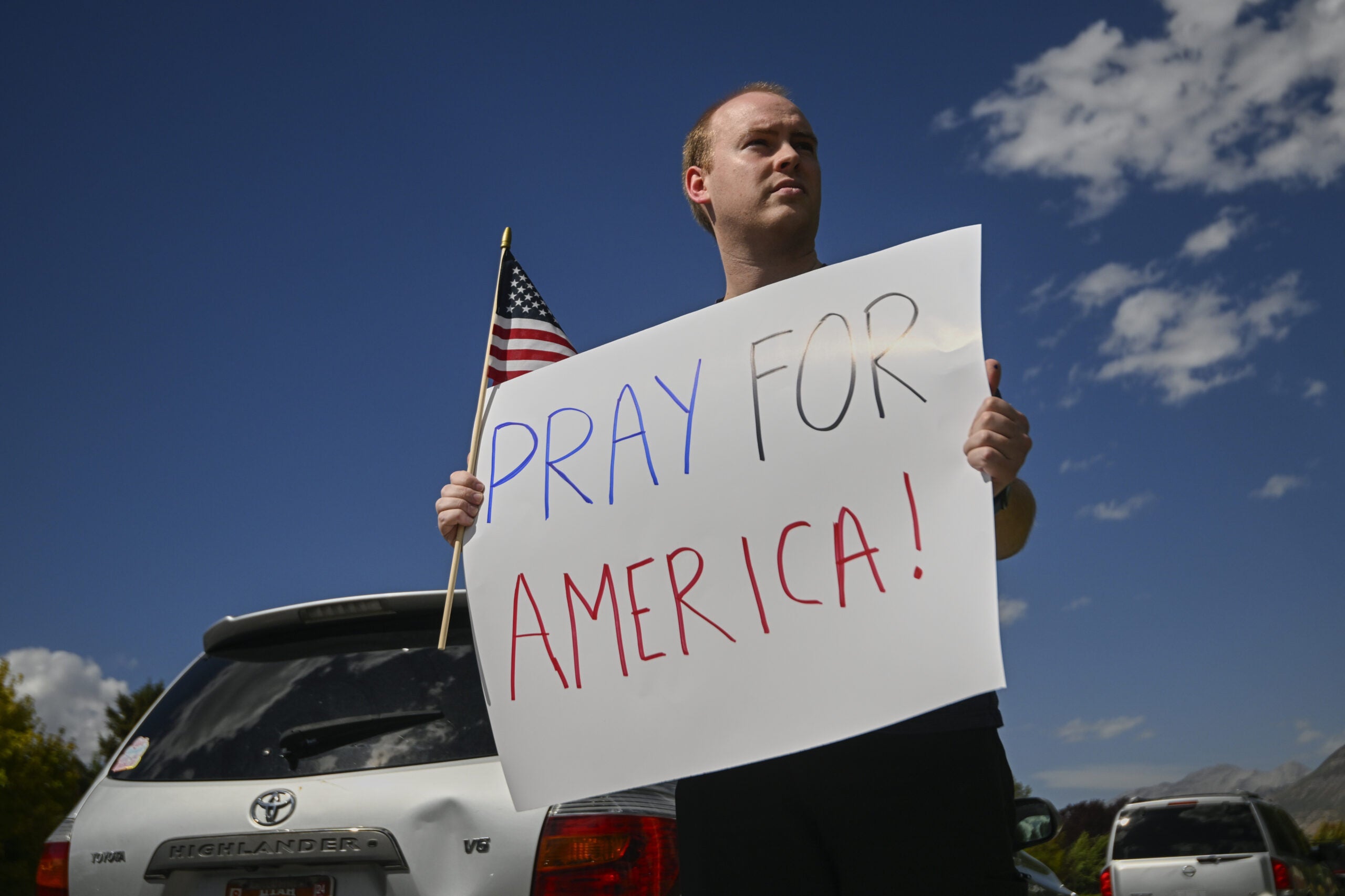 A person stands next to a parked car holding an American flag and a large sign that reads PRAY FOR AMERICA! against a blue sky with some clouds.