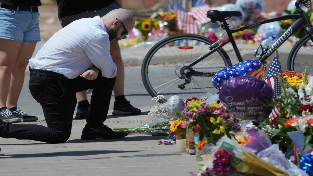 A man kneels and bows his head at a memorial site with flowers, balloons, American flags, and a bicycle, while others pay their respects nearby.