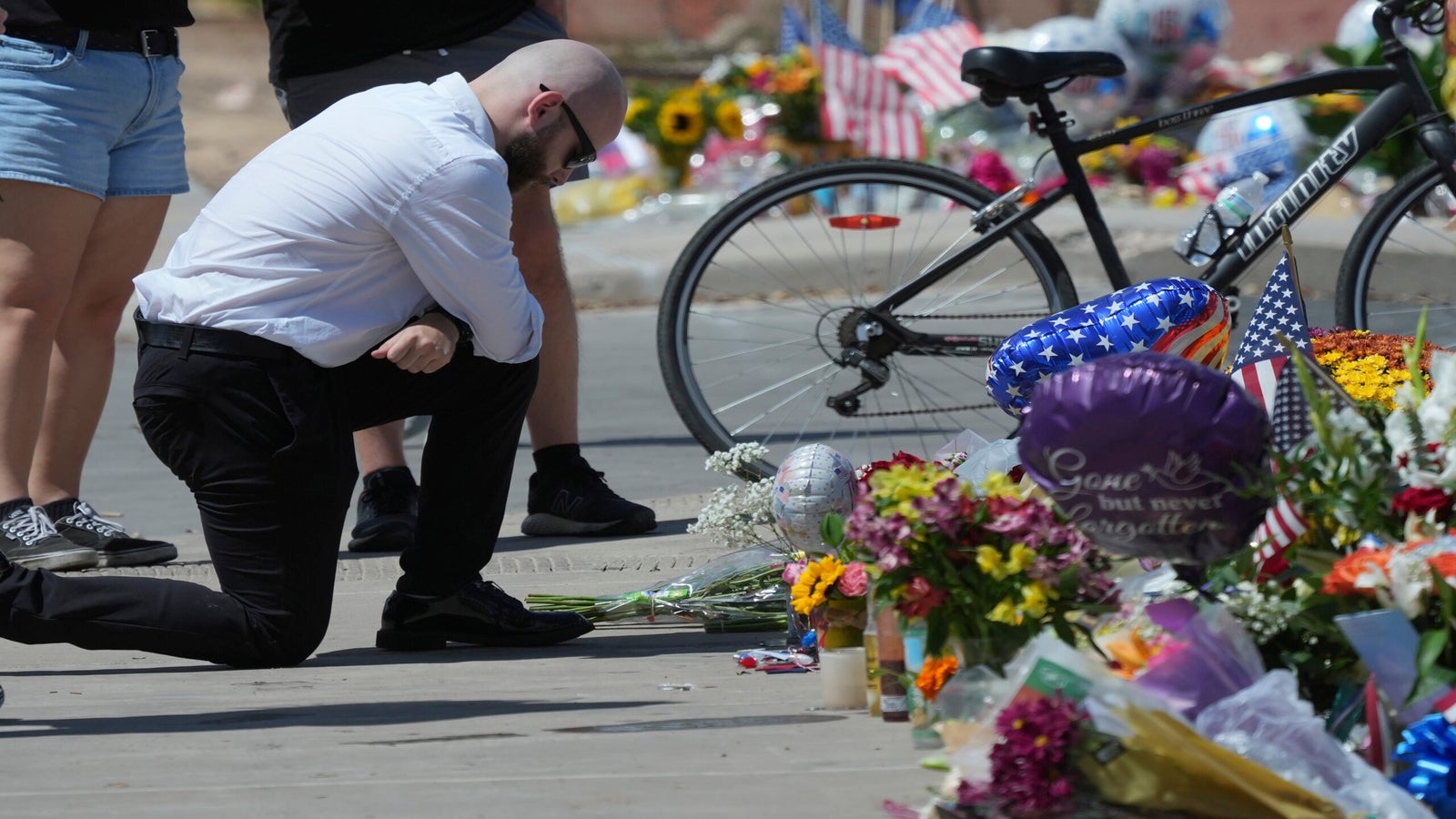 A man kneels and bows his head at a memorial site with flowers, balloons, American flags, and a bicycle, while others pay their respects nearby.