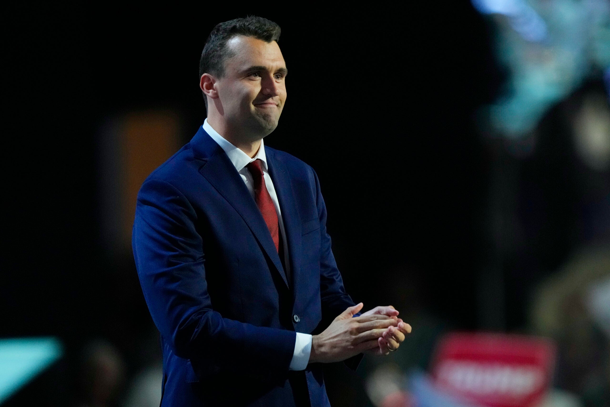 A man in a dark blue suit and red tie stands on stage, clasping his hands, with a dark background and blurred audience.