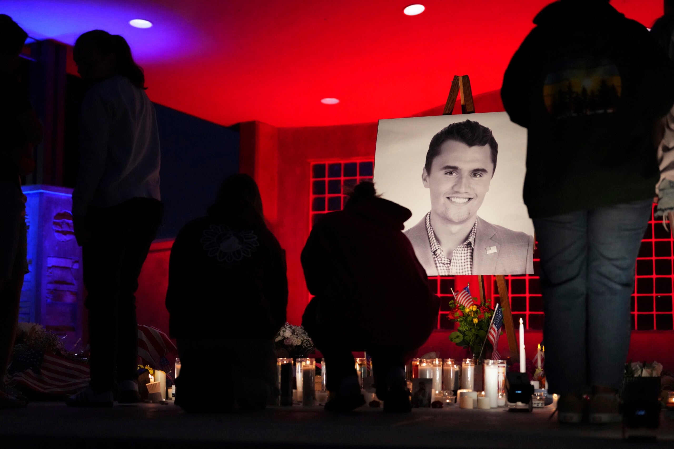 People gather at a candlelight vigil with a large portrait of a man displayed, surrounded by lit candles and flowers under red and blue lighting.
