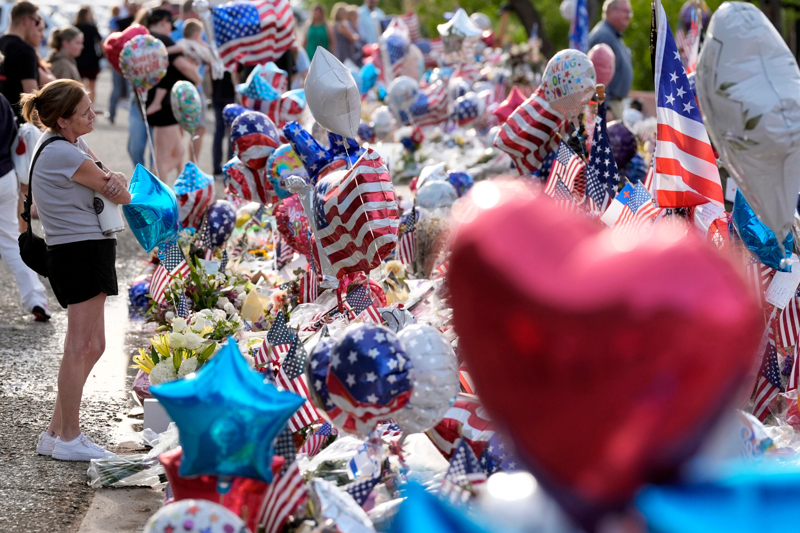 A woman stands among a memorial display of American flags, balloons, and flowers, surrounded by other people.