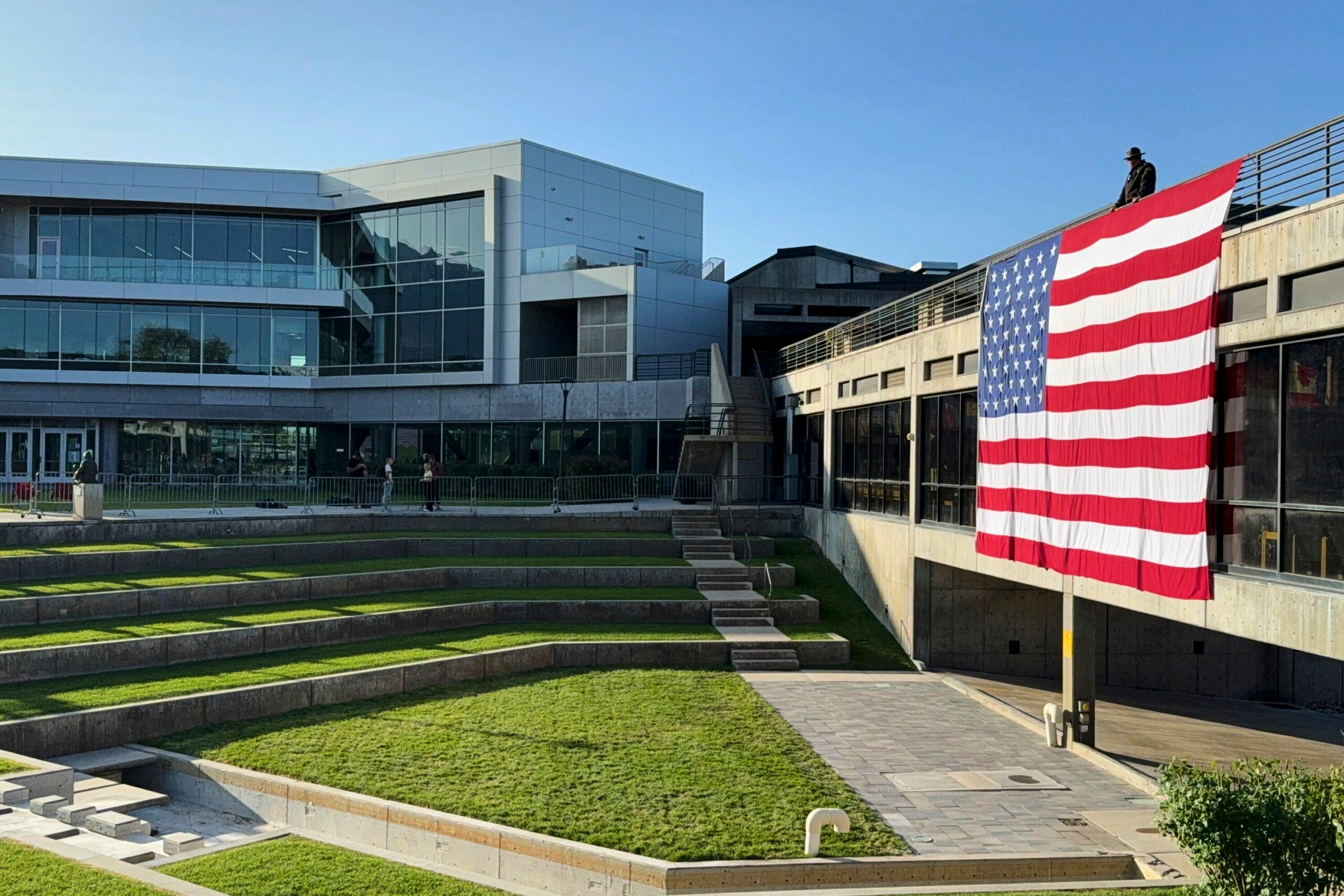 A large American flag hangs from a balcony of a modern building overlooking a stepped grassy amphitheater under a clear sky.