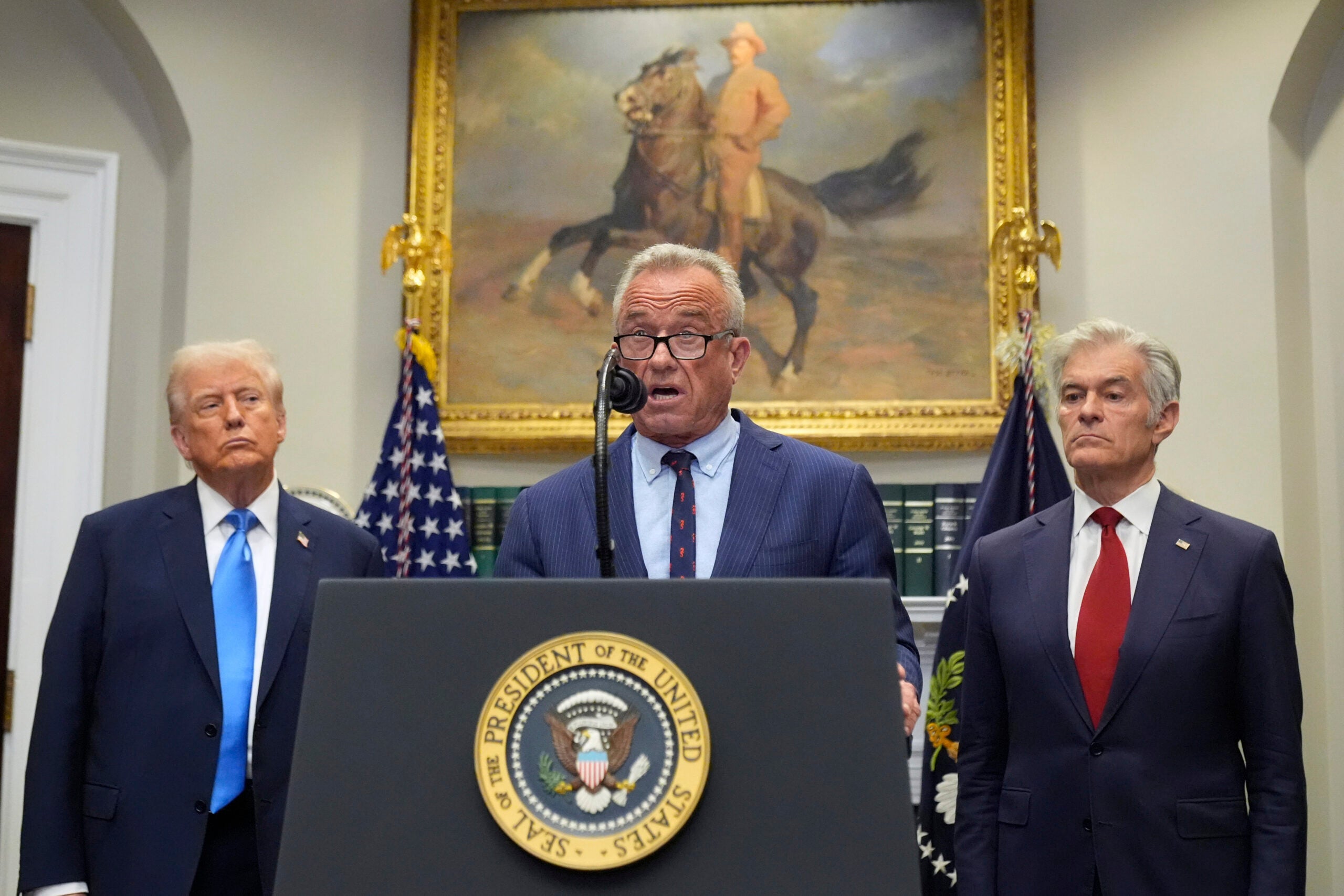 Three men stand at a podium with the Seal of the President of the United States, flanked by flags and a painting of a rider on horseback in the background.