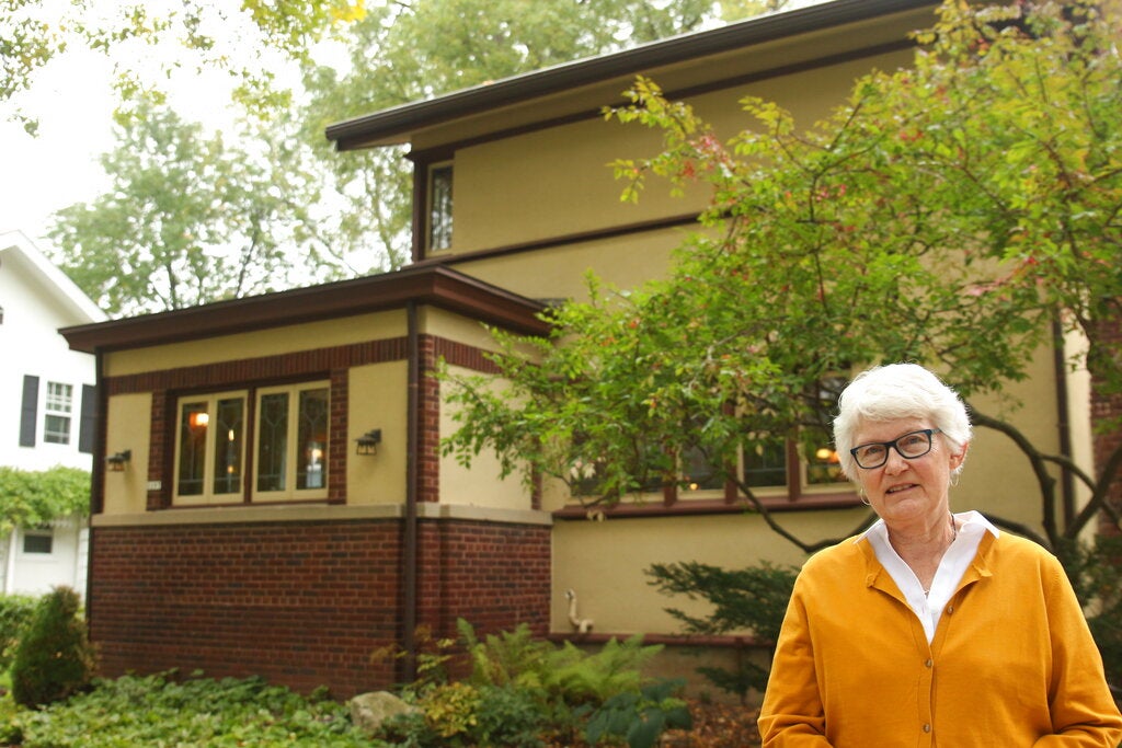 An older woman with short white hair and glasses stands in front of a two-story house with tan and brick exterior, surrounded by greenery.