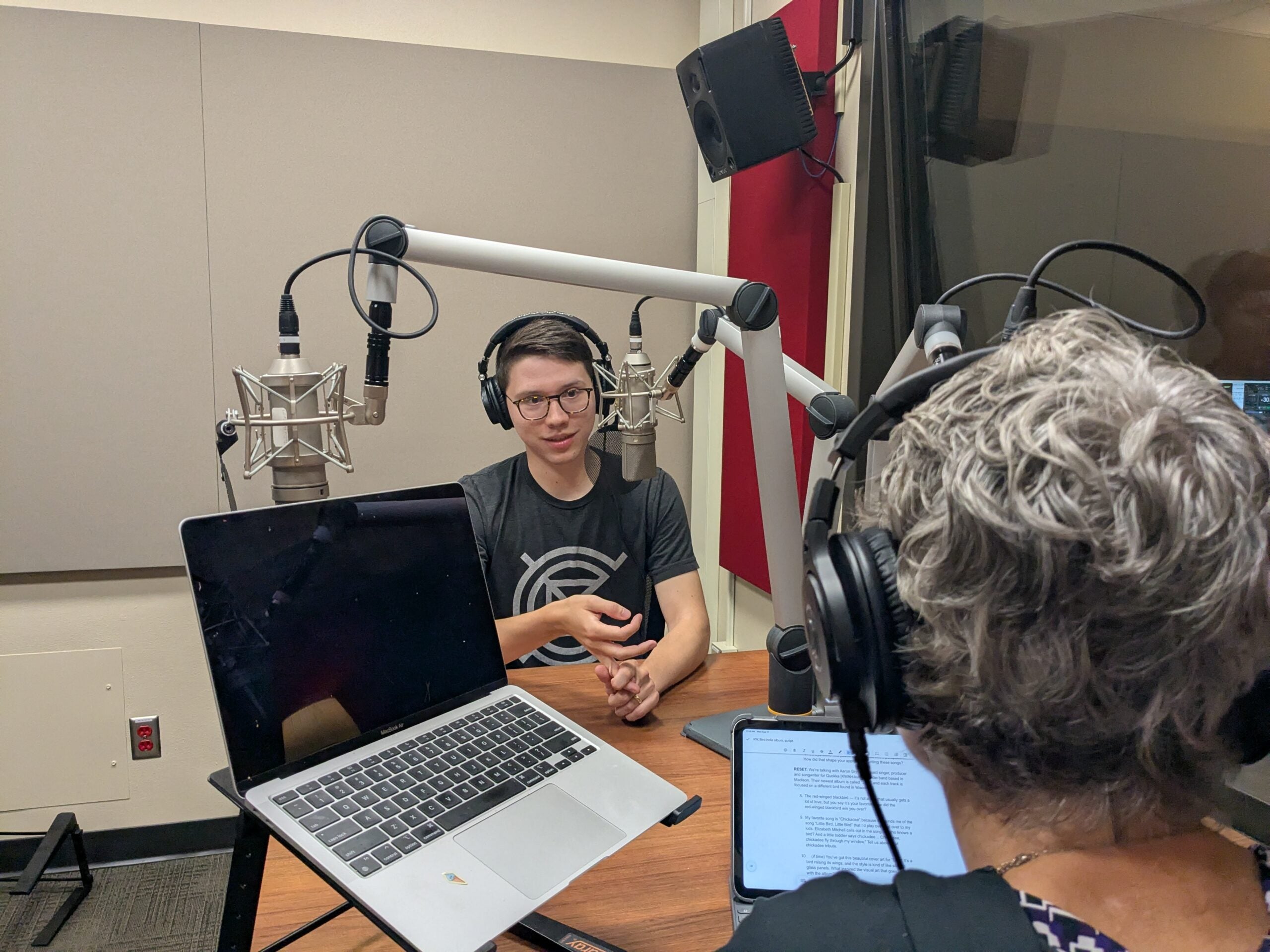 Two people wearing headphones sit at a table in a recording studio with microphones, a laptop, and a tablet in front of them.