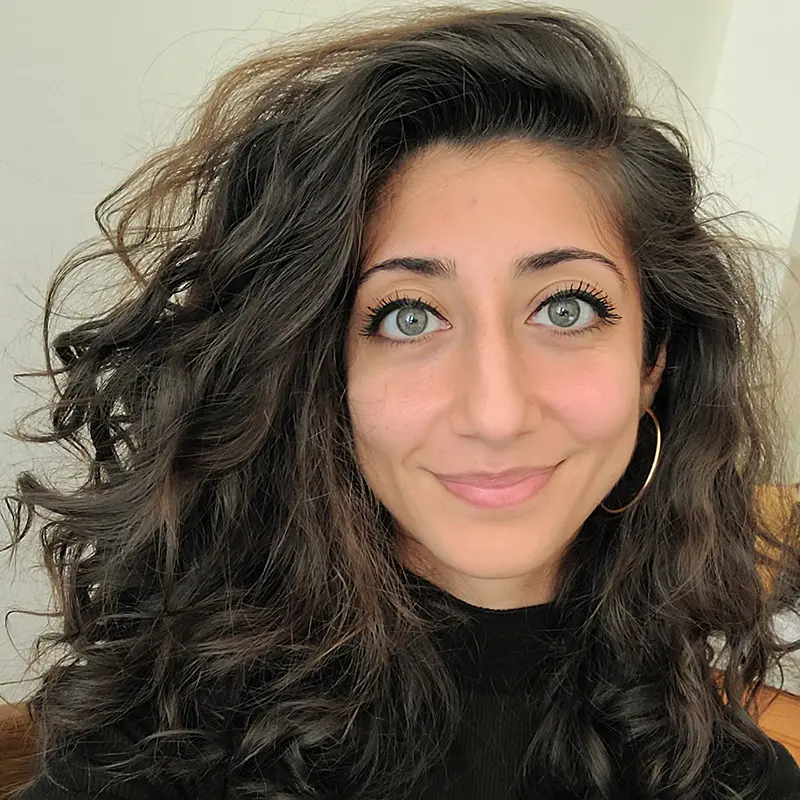 A woman with wavy dark hair, light eyes, and hoop earrings is smiling slightly at the camera. She is wearing a black top and is indoors.