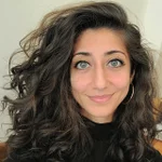 A woman with wavy dark hair, light eyes, and hoop earrings is smiling slightly at the camera. She is wearing a black top and is indoors.