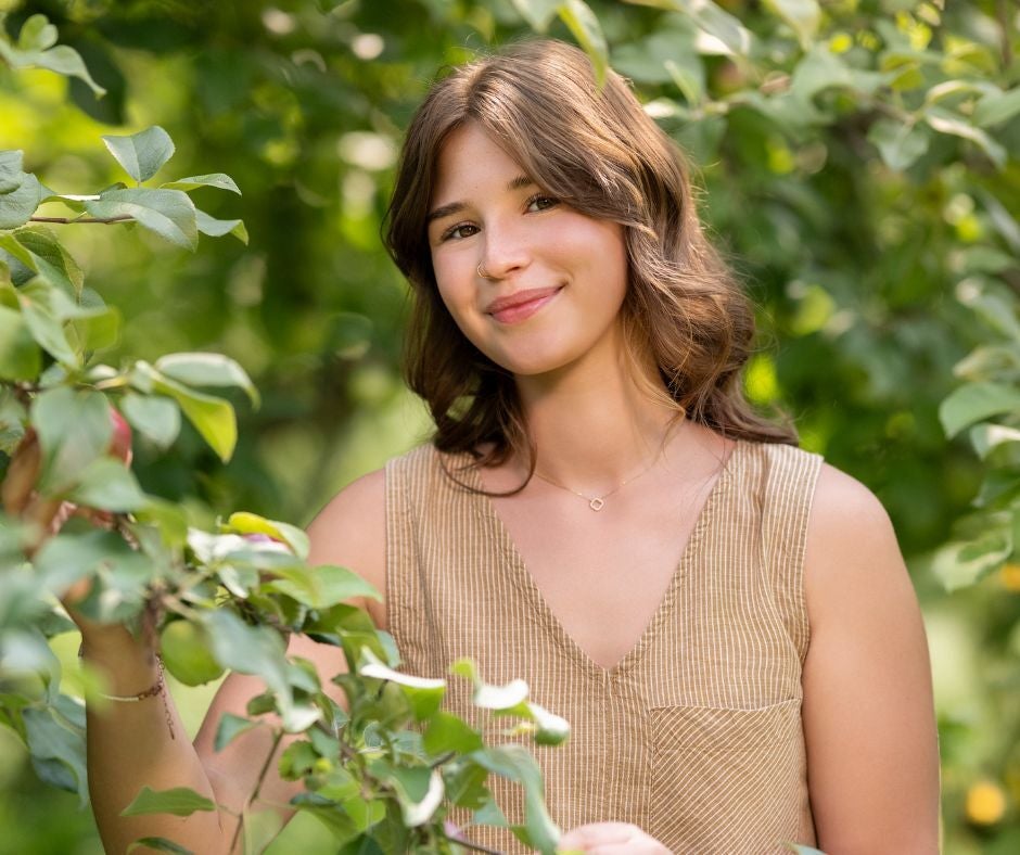 A young woman with brown hair stands outdoors among green leafy branches, wearing a sleeveless beige top and smiling at the camera.