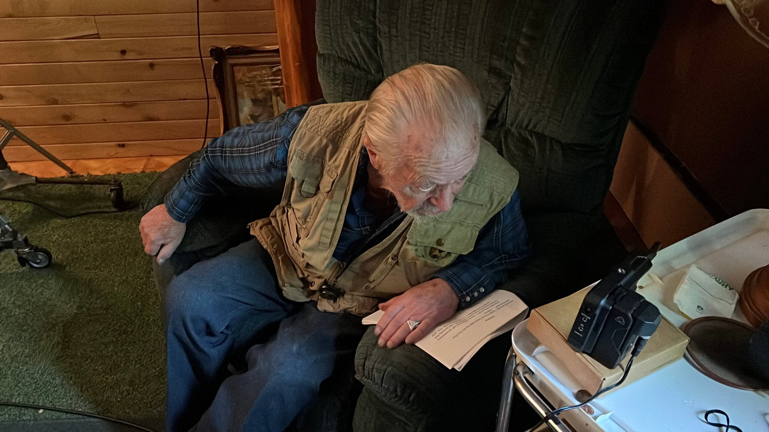 An elderly man with gray hair sits in a recliner, looking down at a sheet of paper on a tray next to him in a dimly lit room.