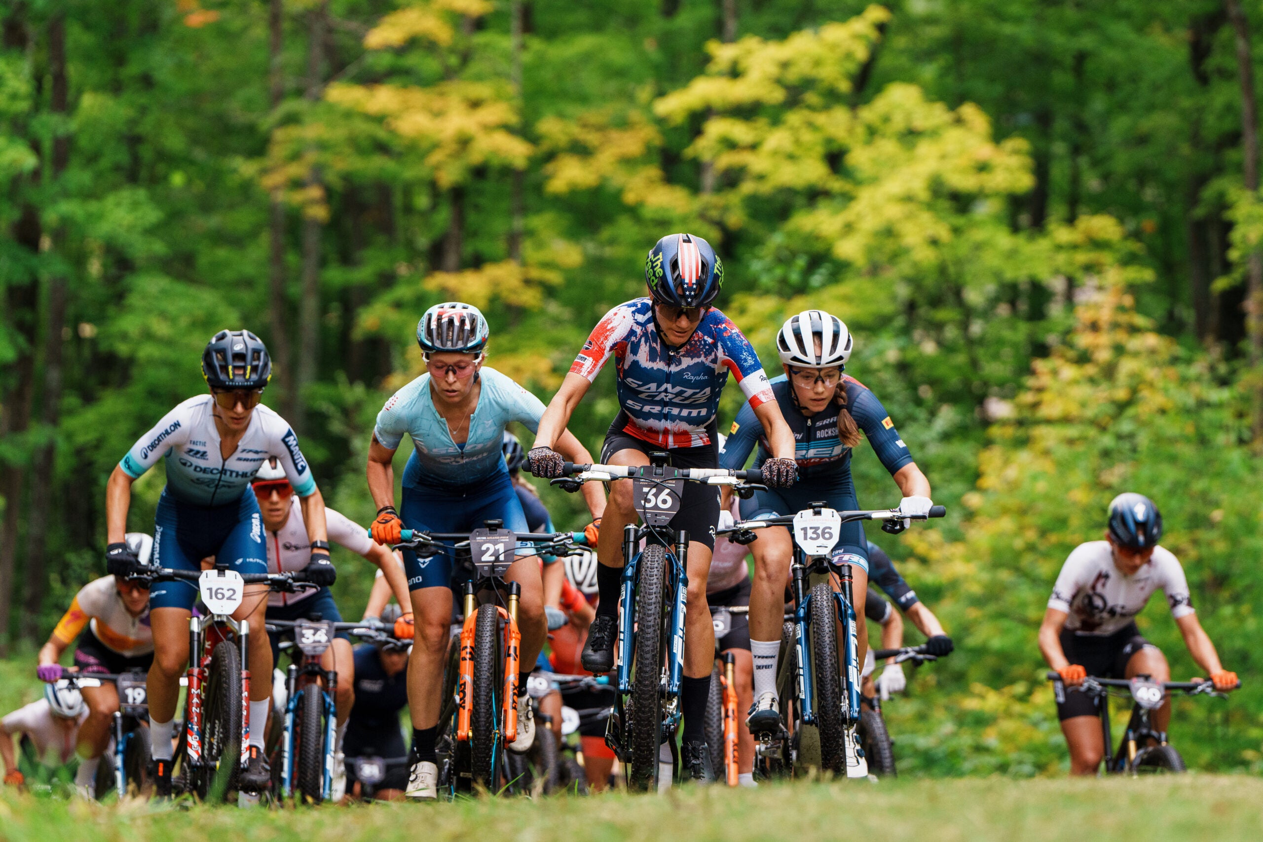 Sekelompok pengendara sepeda berkompetisi dalam perlombaan sepeda gunung melalui kawasan hutan, dengan beberapa pengendara mengenakan kaos dan helm bernomor.