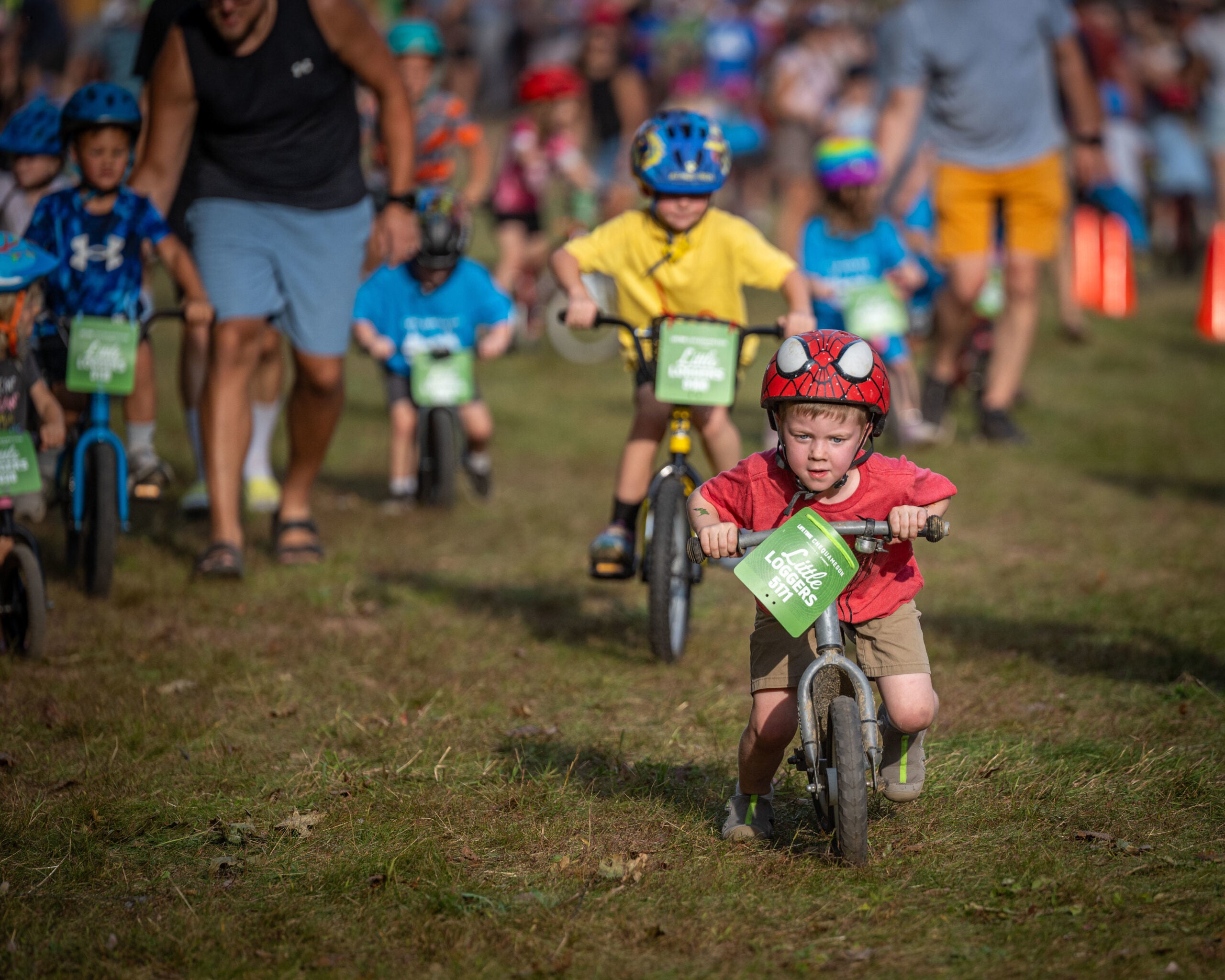 Sekelompok anak kecil yang mengenakan helm mengendarai sepeda keseimbangan dalam perlombaan luar ruangan berumput, dengan orang dewasa dan lebih banyak anak-anak di latar belakang.