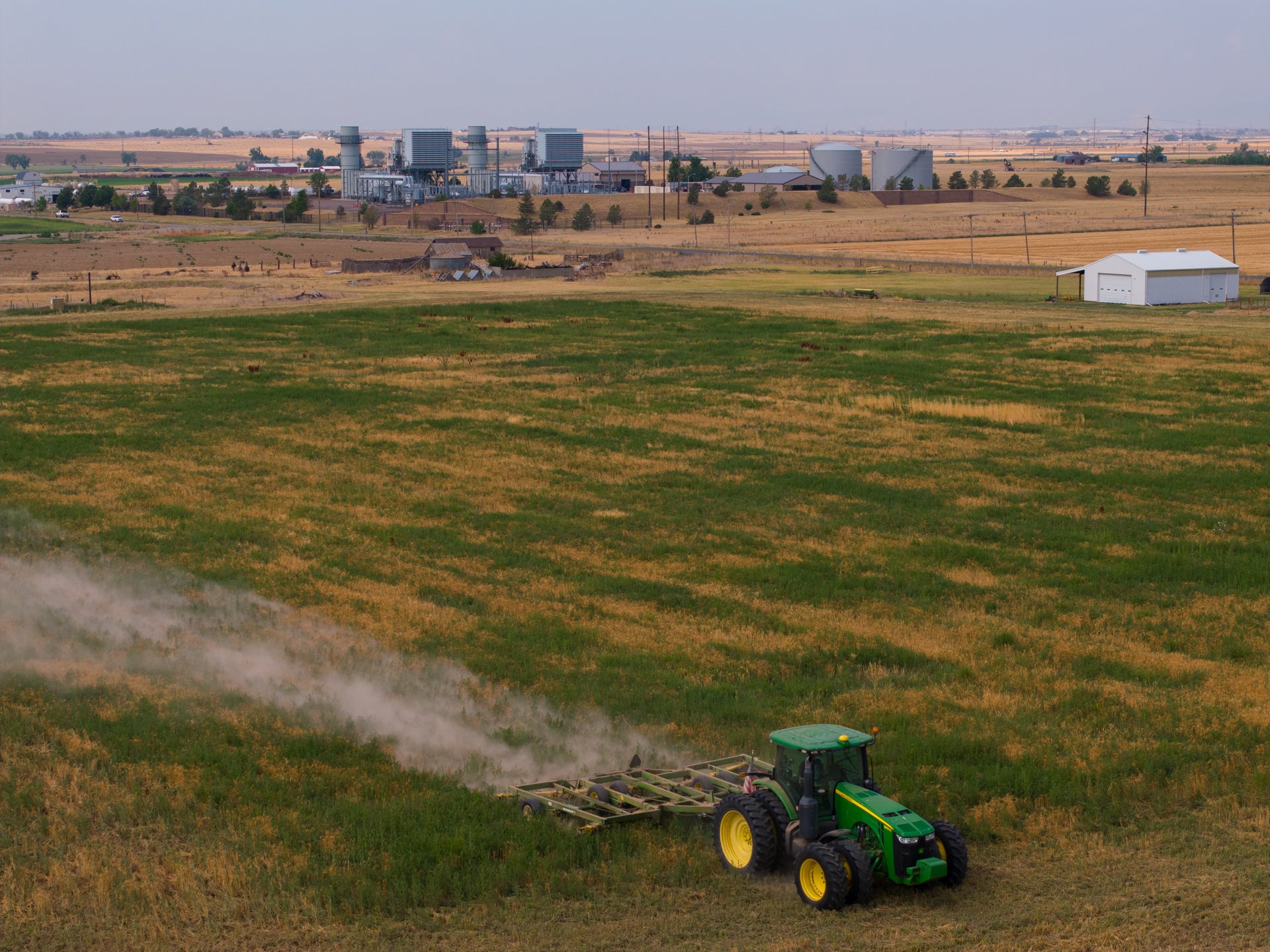 A green tractor tills a field, raising dust, with industrial buildings and storage tanks visible in the background.
