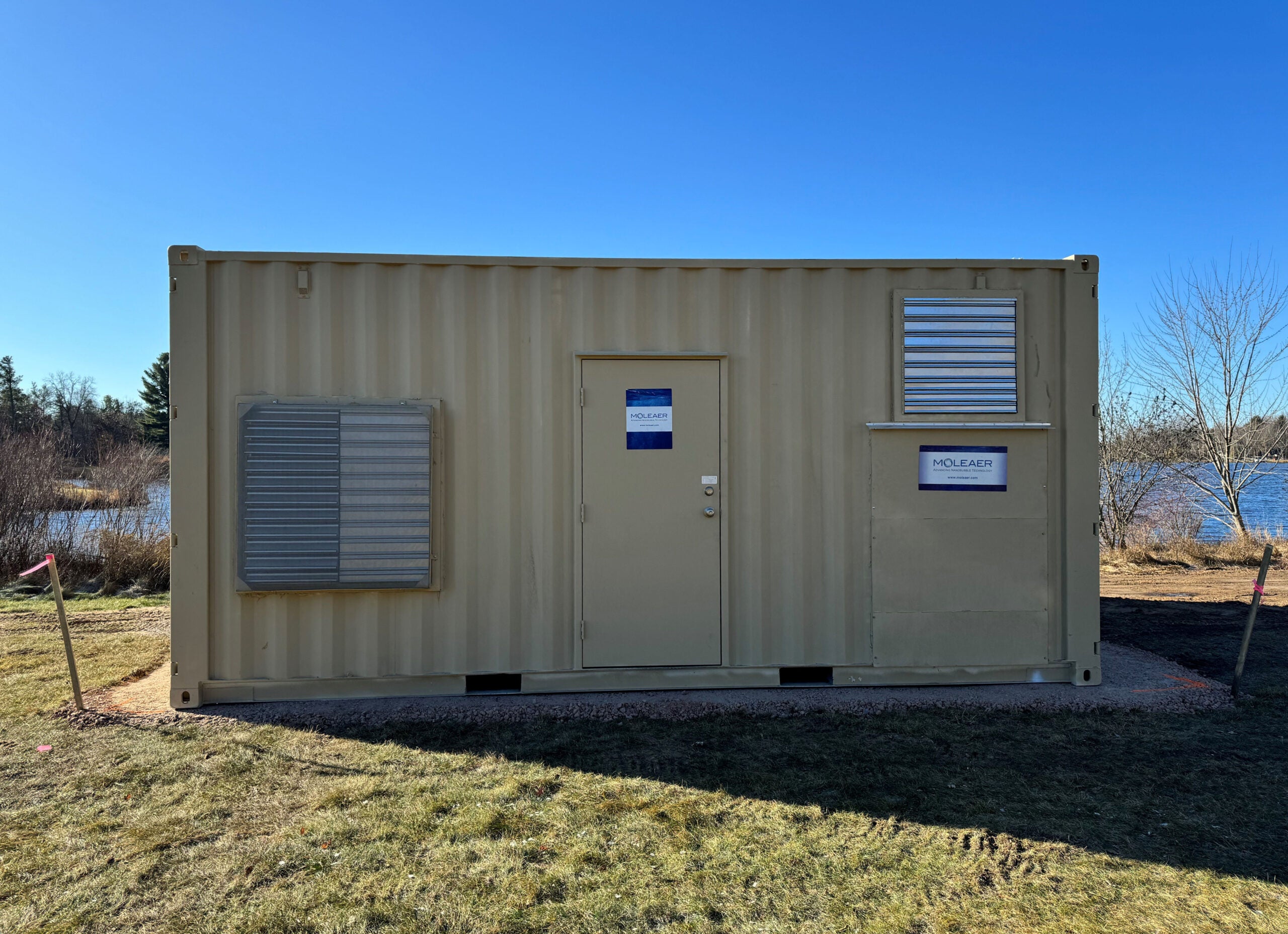 A beige shipping container with a door and two vented windows sits on grass near a pond, under a clear blue sky. Signs on the door and wall indicate it is a research facility.